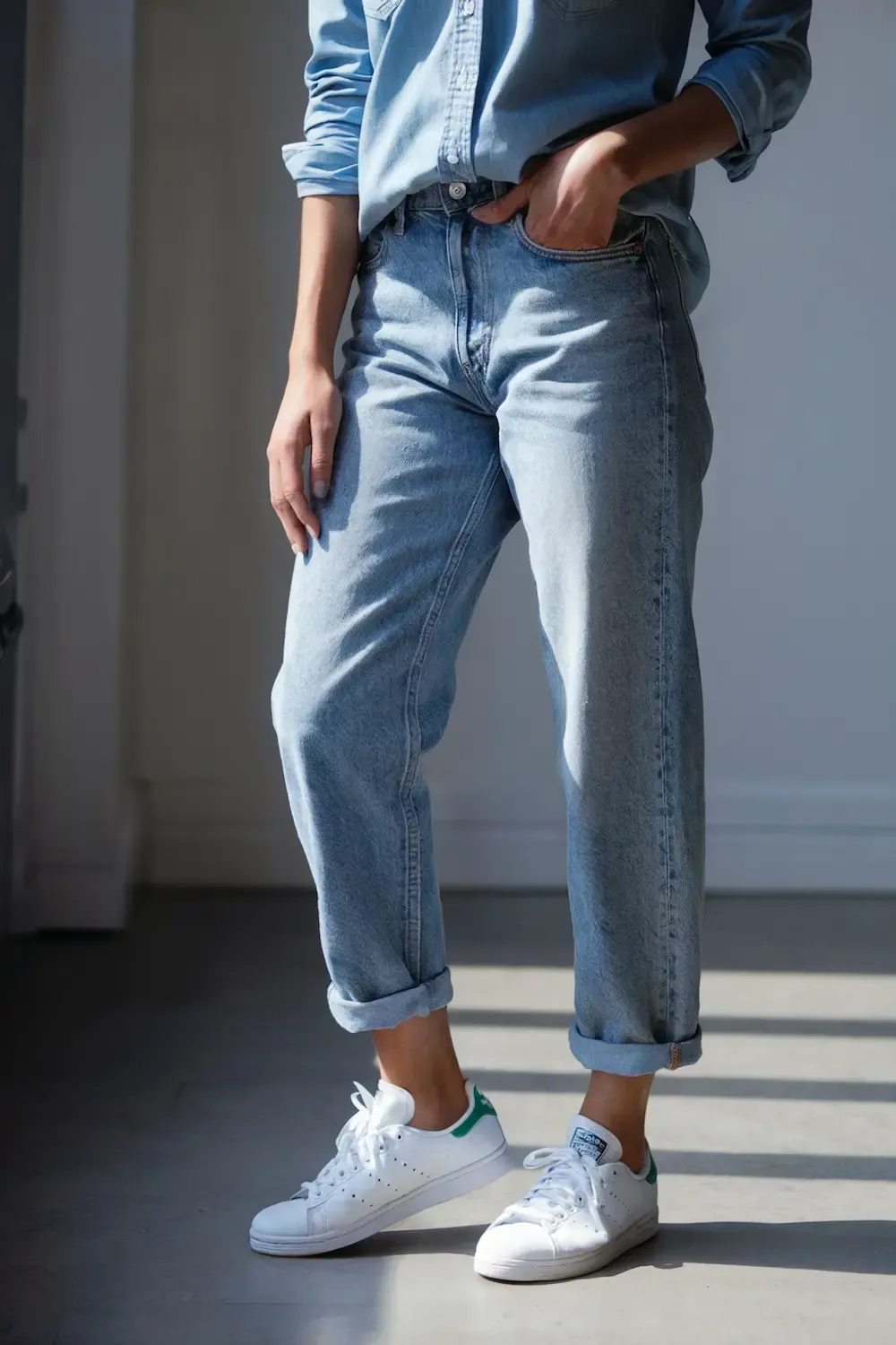 Woman wearing rolled light wash straight leg jeans with white and green Stan Smith sneakers and chambray button-down shirt in natural lighting