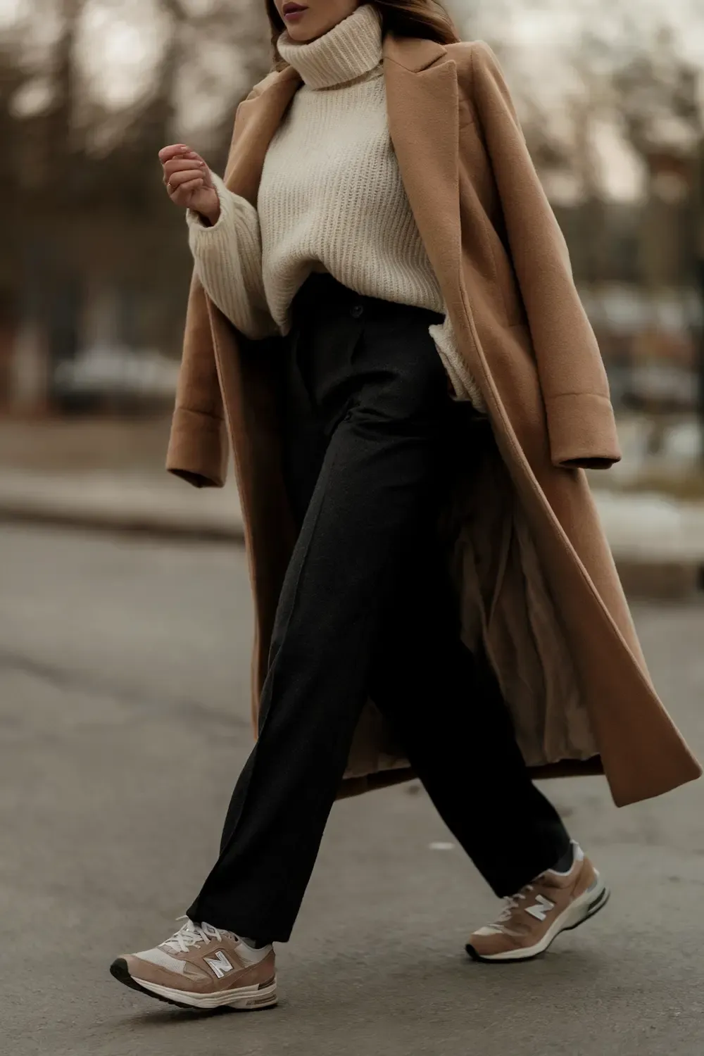 Woman wearing camel coat, cream turtleneck sweater, black trousers, and beige New Balance sneakers walking on urban street, demonstrating luxe casual winter layering