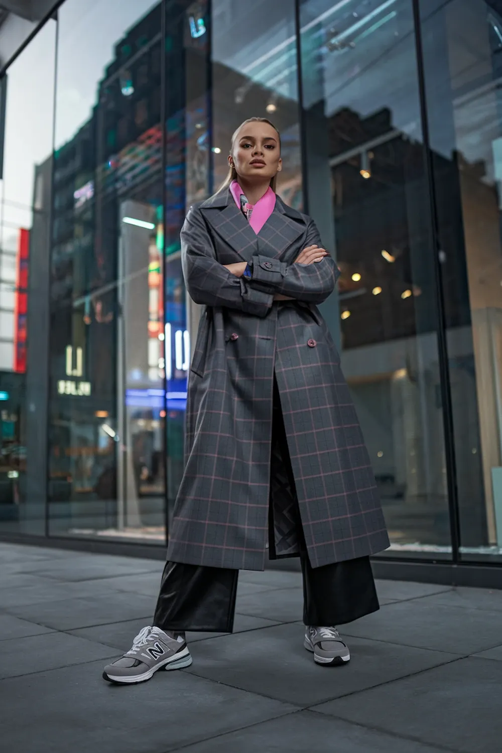 Model wearing dark gray plaid trench coat with pink collar detail paired with gray New Balance 574 sneakers in urban city setting, demonstrating sophisticated street style against a modern glass building backdrop.
