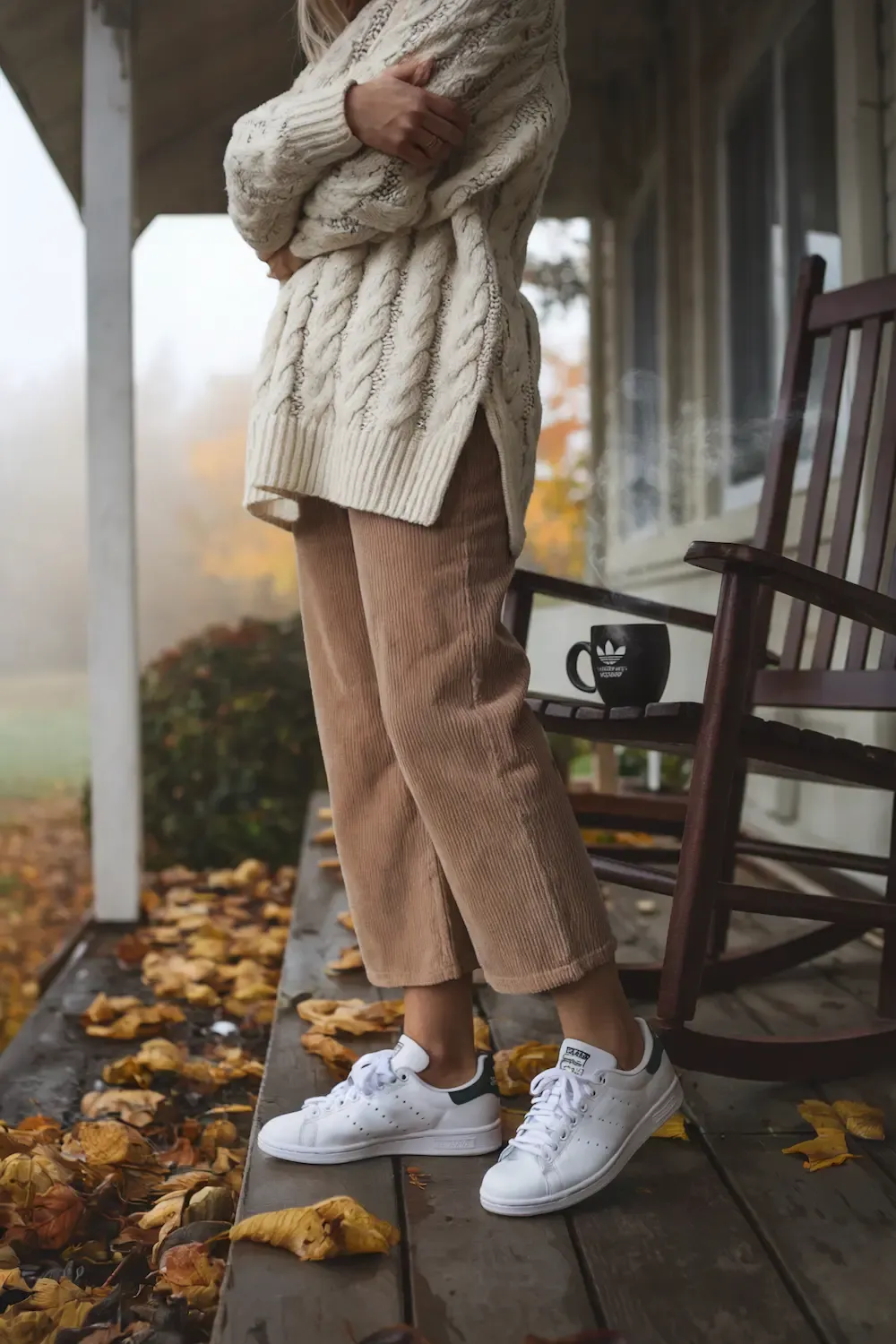 Cream cable-knit oversized sweater paired with beige corduroy pants and white Adidas Stan Smith sneakers for comfortable work-from-home style on a cozy porch setting with autumn leaves