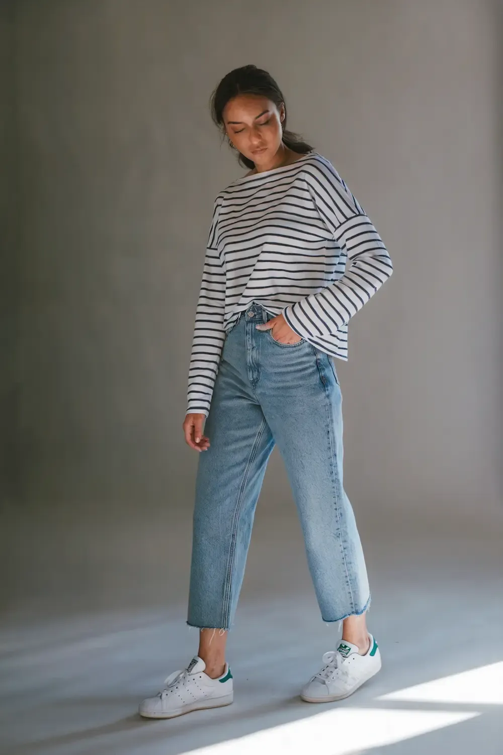 Woman in striped boat neck top paired with straight-cut light denim jeans and classic Stan Smith sneakers, standing in studio lighting