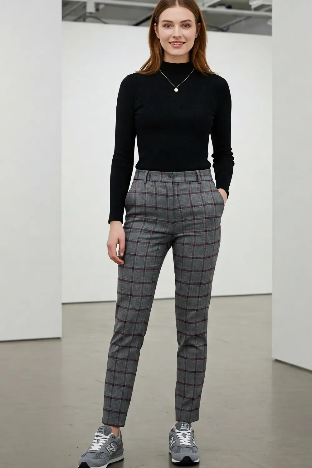 Woman wearing New Balance 990v6 grey sneakers styled with a black fitted mock neck top and grey plaid tailored trousers in a minimal studio setting