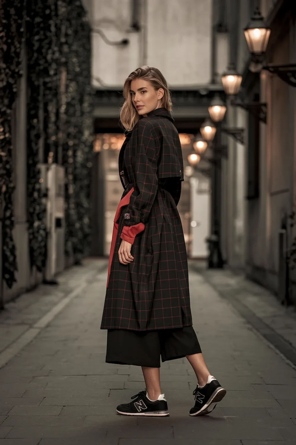 Model wearing dark brown plaid trench coat styled with black New Balance 574 sneakers in historic alleyway with ambient lighting, showcasing elegant evening smart-casual styling with wide-leg black pants.