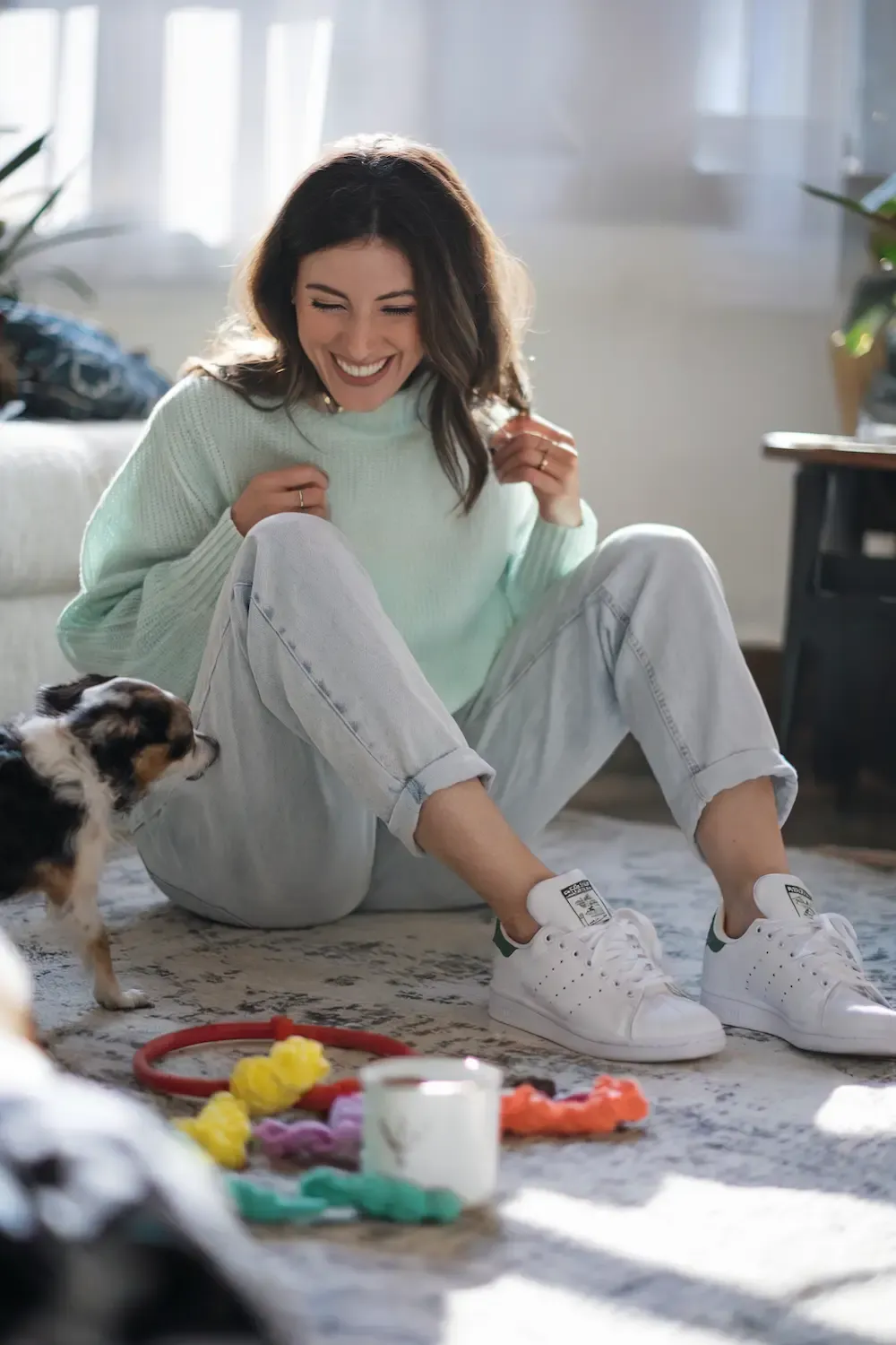 Woman in mint green sweater and light wash jeans wearing white Stan Smith sneakers playing with small dog on patterned rug in bright living room - casual work-from-home outfit for active days