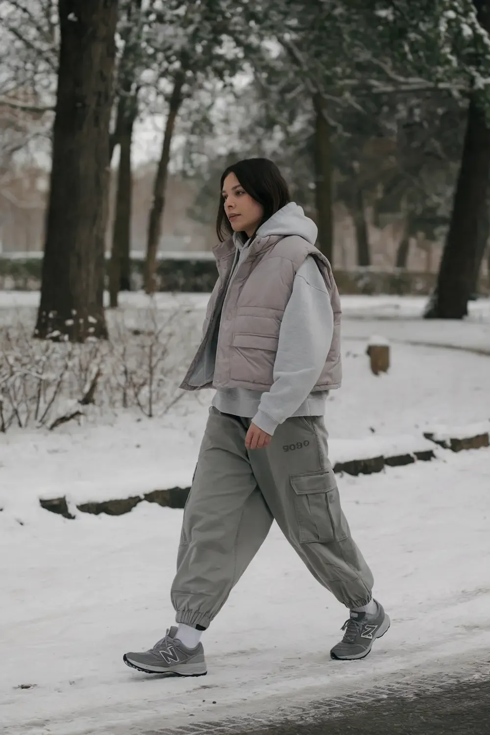 Woman in grey puffer vest, light grey hoodie, olive cargo pants, and grey New Balance sneakers walking in snowy park setting, showcasing modern athleisure style