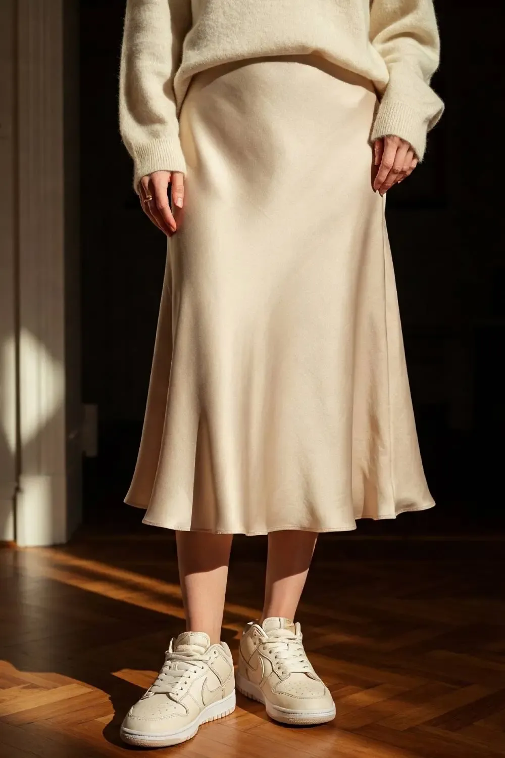 Close-up of a woman's outfit showing the texture contrast between a cream cashmere sweater, champagne silk skirt, and white Nike Dunk Lows.