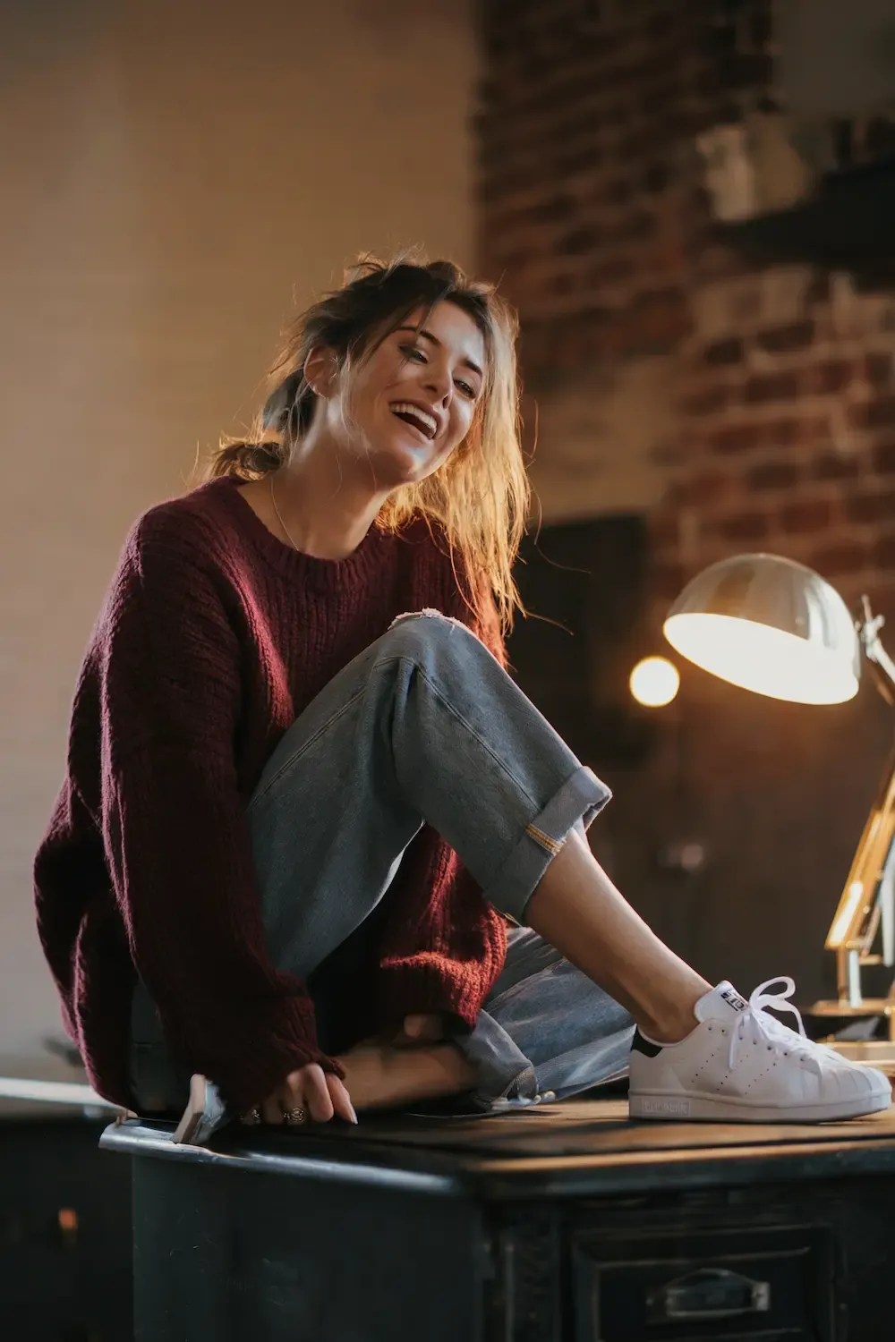 Burgundy ribbed sweater styled with light wash jeans and white Stan Smith sneakers for a warm work-from-home look in a well-lit home office with brick accent wall