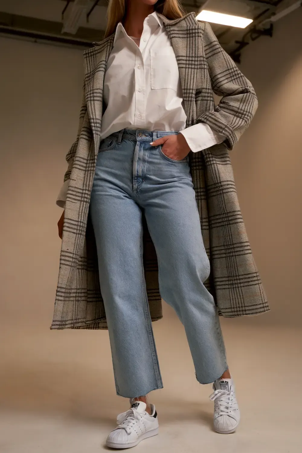Woman wearing check pattern coat with white button-down shirt, light wash straight jeans, and Stan Smith sneakers for a preppy casual style