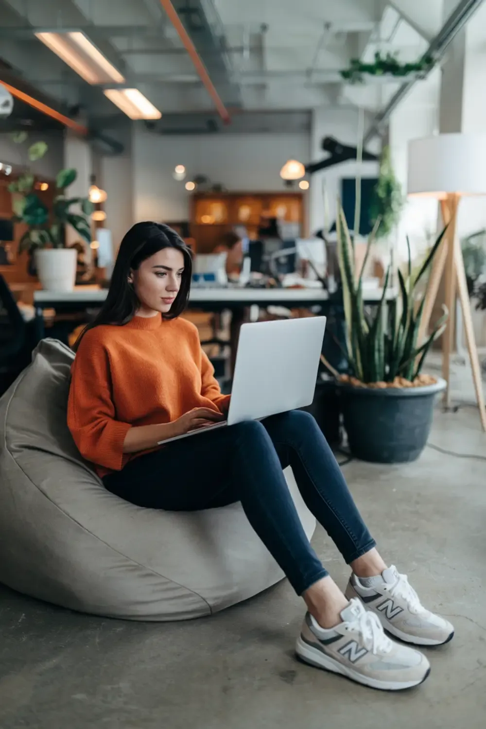 Woman in bright orange sweater working on laptop in modern co-working space wearing white New Balance sneakers, representing comfortable creative professional style.