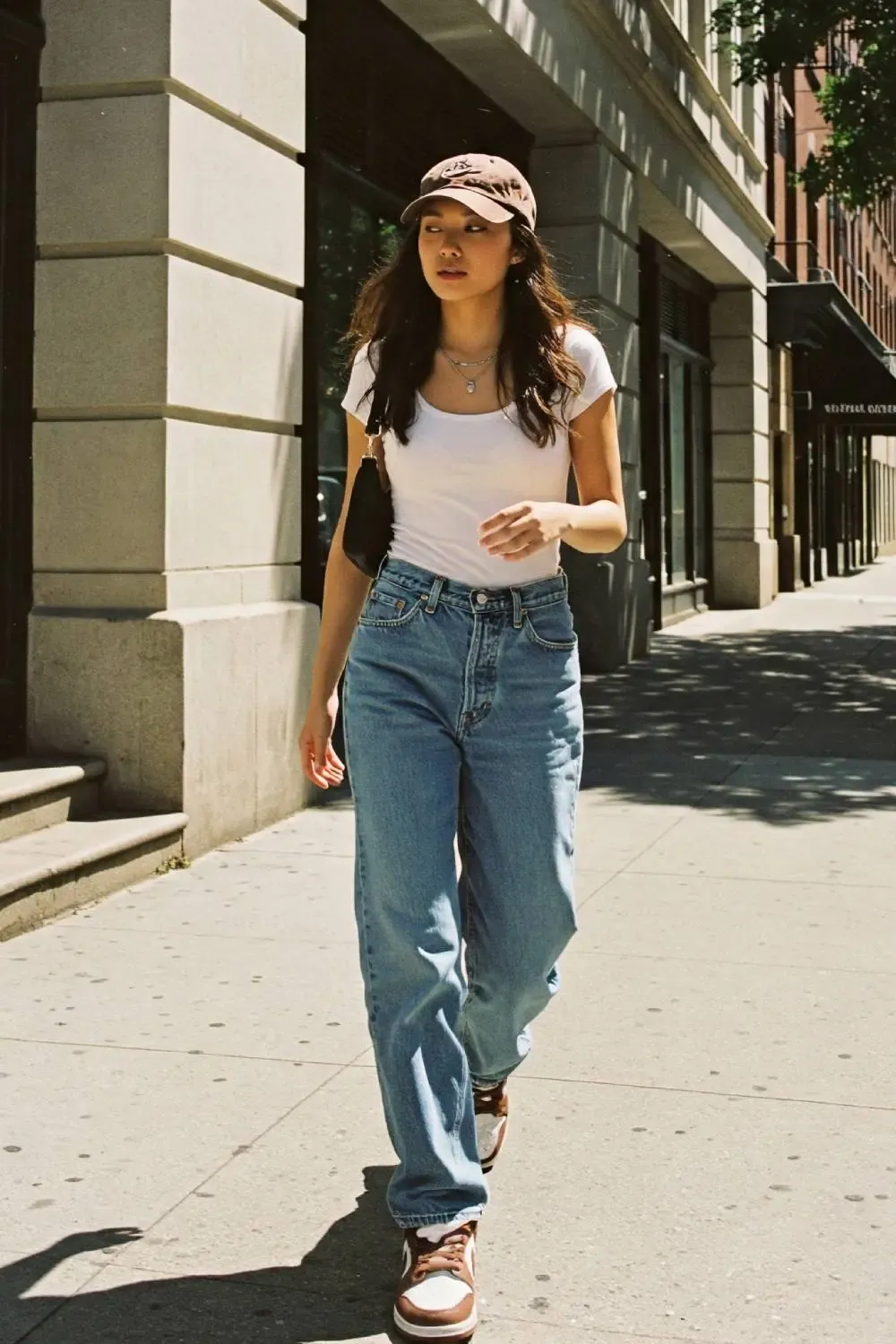 Woman in light-wash baggy jeans and 'Medium Curry' Nike Dunk Lows, paired with a white baby tee, baseball cap, and shoulder bag, epitomizing street style.
