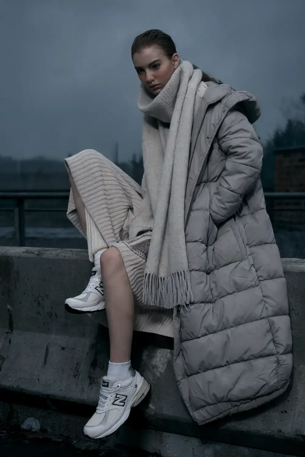 Woman wearing long grey puffer coat, cream knit dress, and white New Balance sneakers sitting on concrete ledge at dusk, showing minimalist winter style