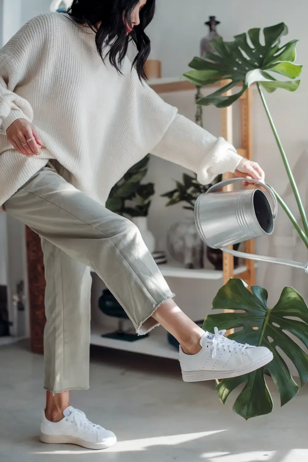 Woman in cream oversized sweater and light wash cropped pants watering houseplants while wearing white sneakers - multitasking work-from-home outfit combining comfort and functionality