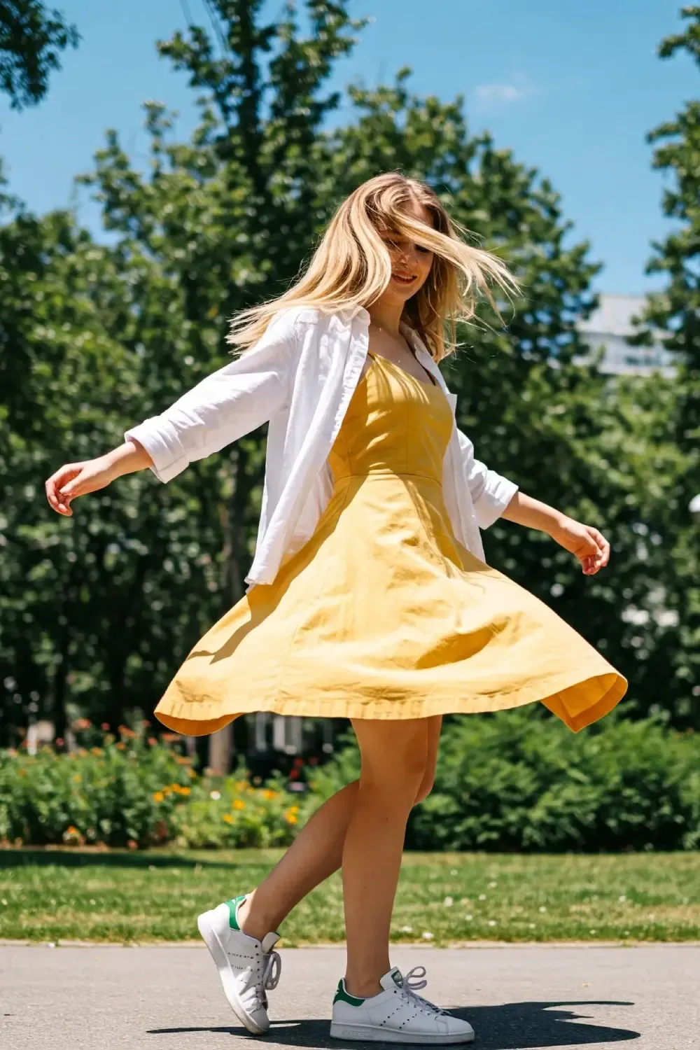 Woman twirling in bright yellow cotton sundress layered with white button-up shirt and Adidas Stan Smith sneakers in a sunny park setting