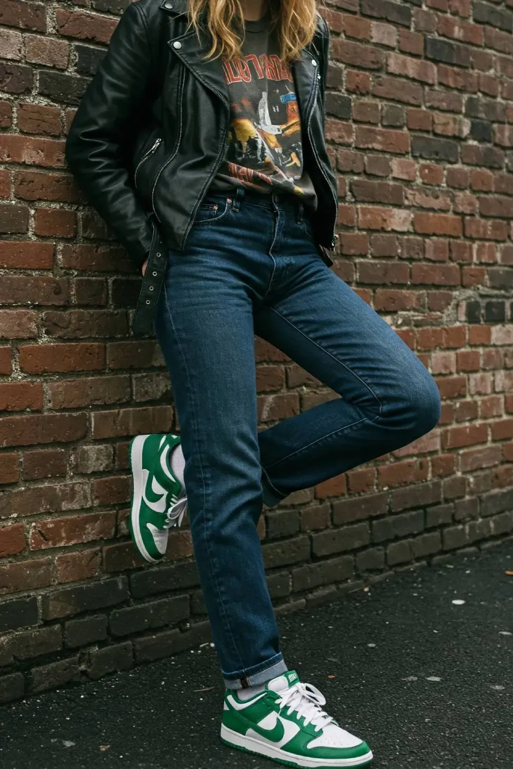 Person in dark wash straight-leg cuffed jeans, 'Brazil' Nike Dunk Lows, a band tee, and leather jacket, creating an edgy look.