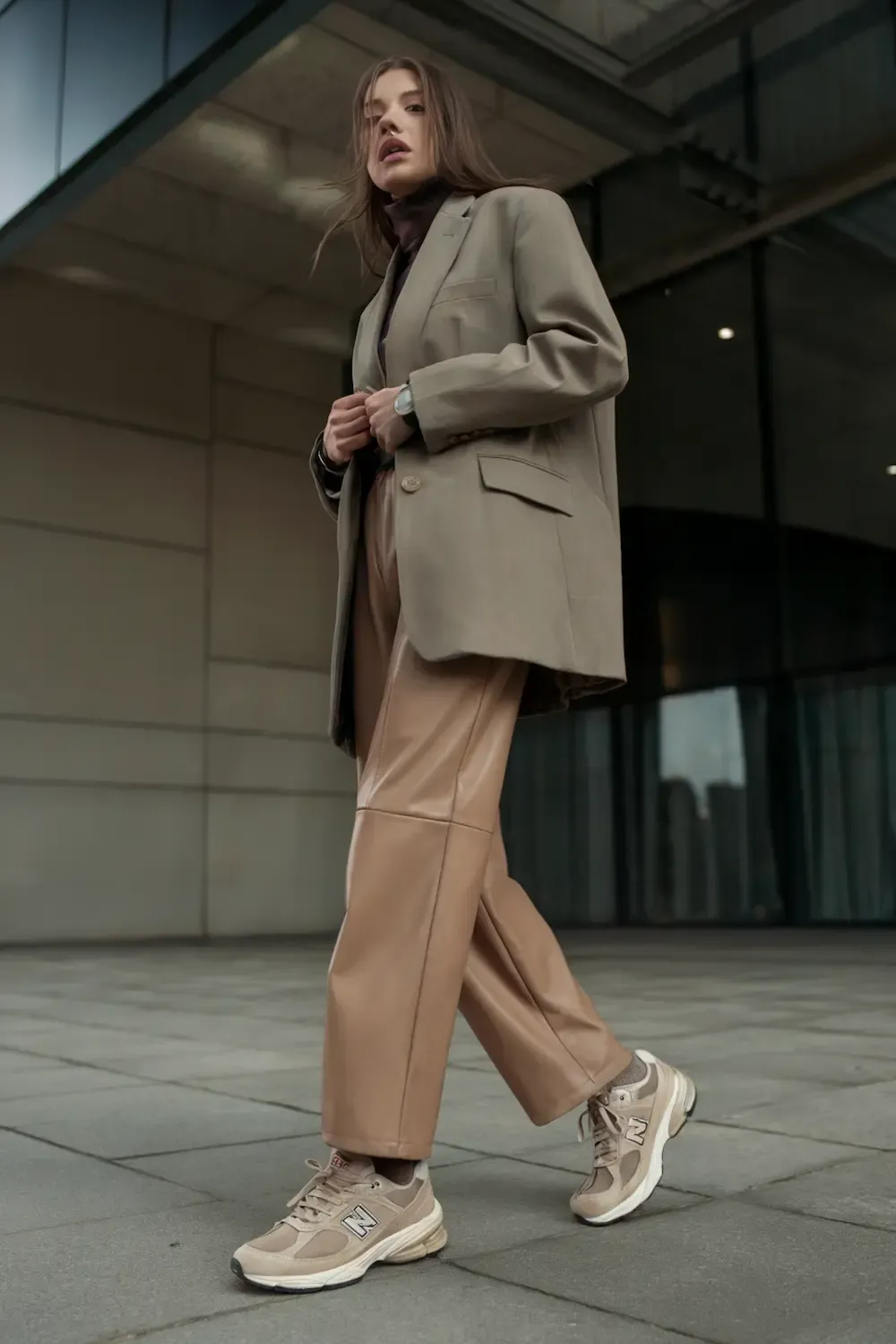 Woman in beige blazer, dark turtleneck, camel leather pants, and beige New Balance sneakers against modern architecture, demonstrating elevated casual workwear
