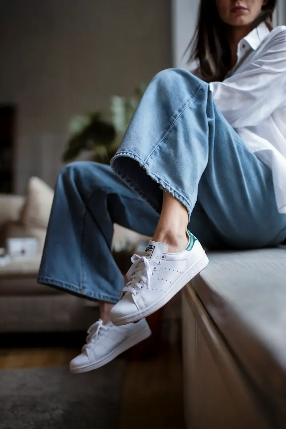 Woman sitting casually with white oversized button-down, light blue wide-leg jeans and white Stan Smith sneakers with green detail in home setting
