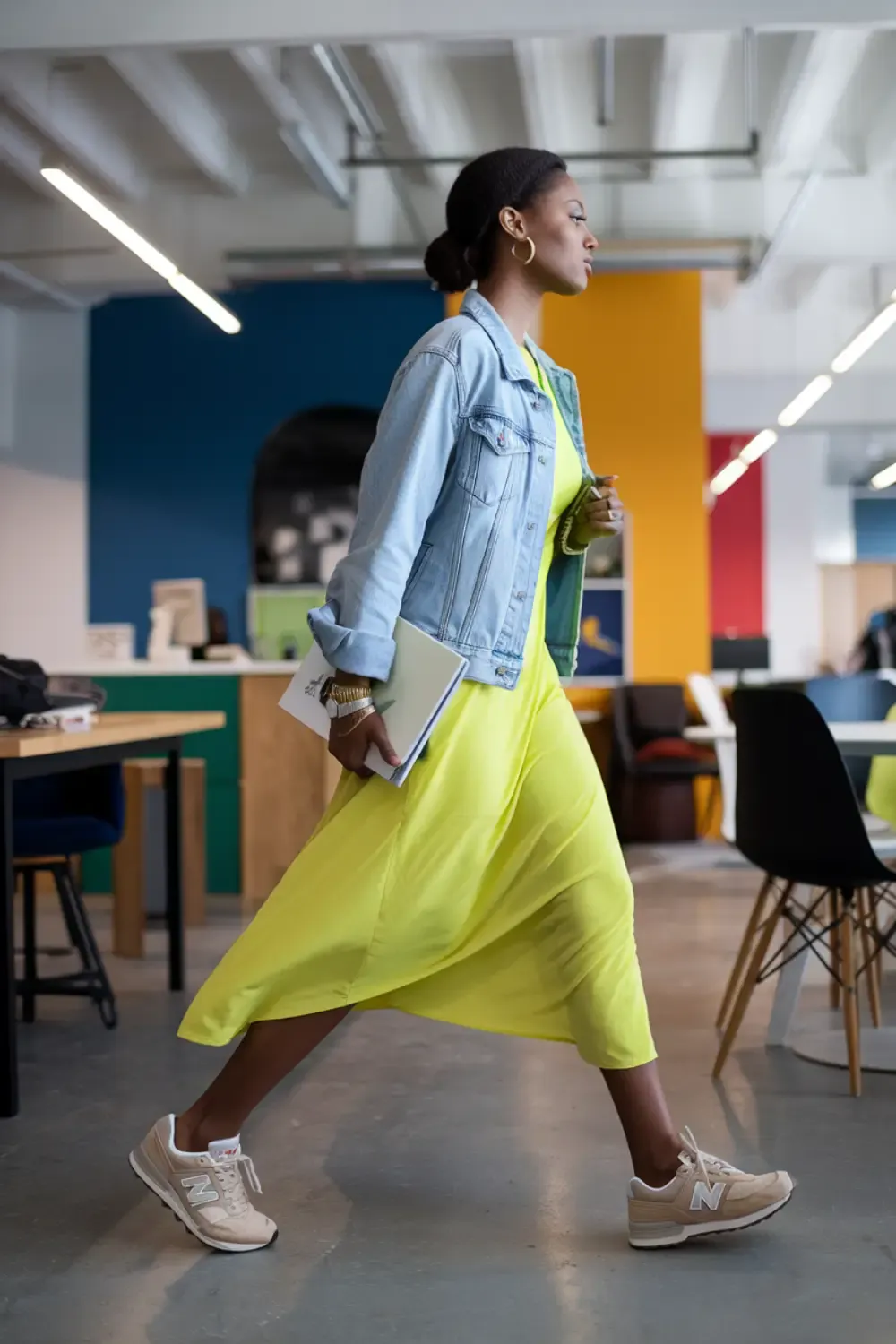 Woman in light denim jacket and yellow dress walking through colorful office space with neutral New Balance sneakers, perfect for business casual environments.