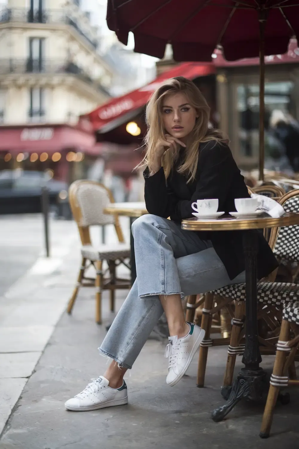 Woman at Parisian café wearing black top, light wash cropped jeans, and white Stan Smith sneakers showcasing effortless European style