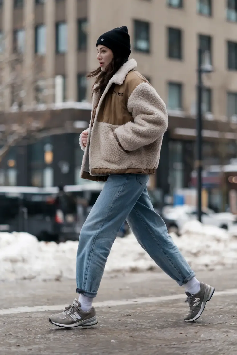 Woman wearing beige sherpa jacket, black beanie, relaxed-fit jeans, and grey New Balance sneakers walking in urban winter setting, showing casual weekend style
