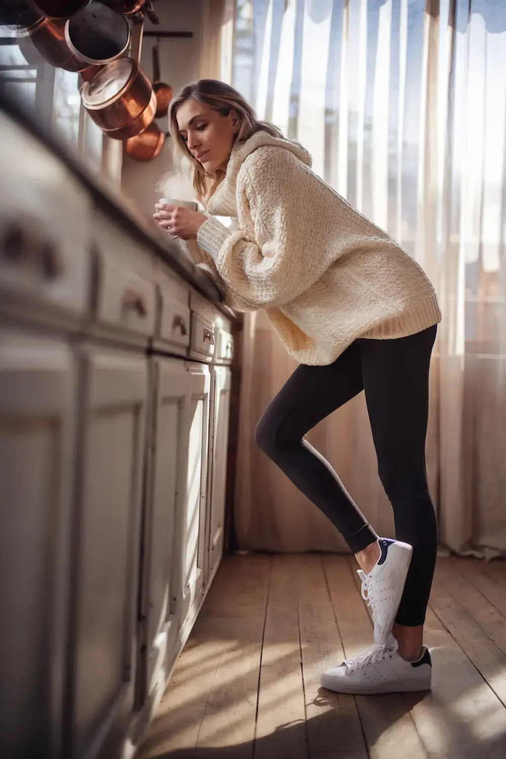 Woman in cream oversized sweater and black leggings with white sneakers enjoying coffee in sunny kitchen - comfortable work-from-home outfit perfect for kitchen workspace