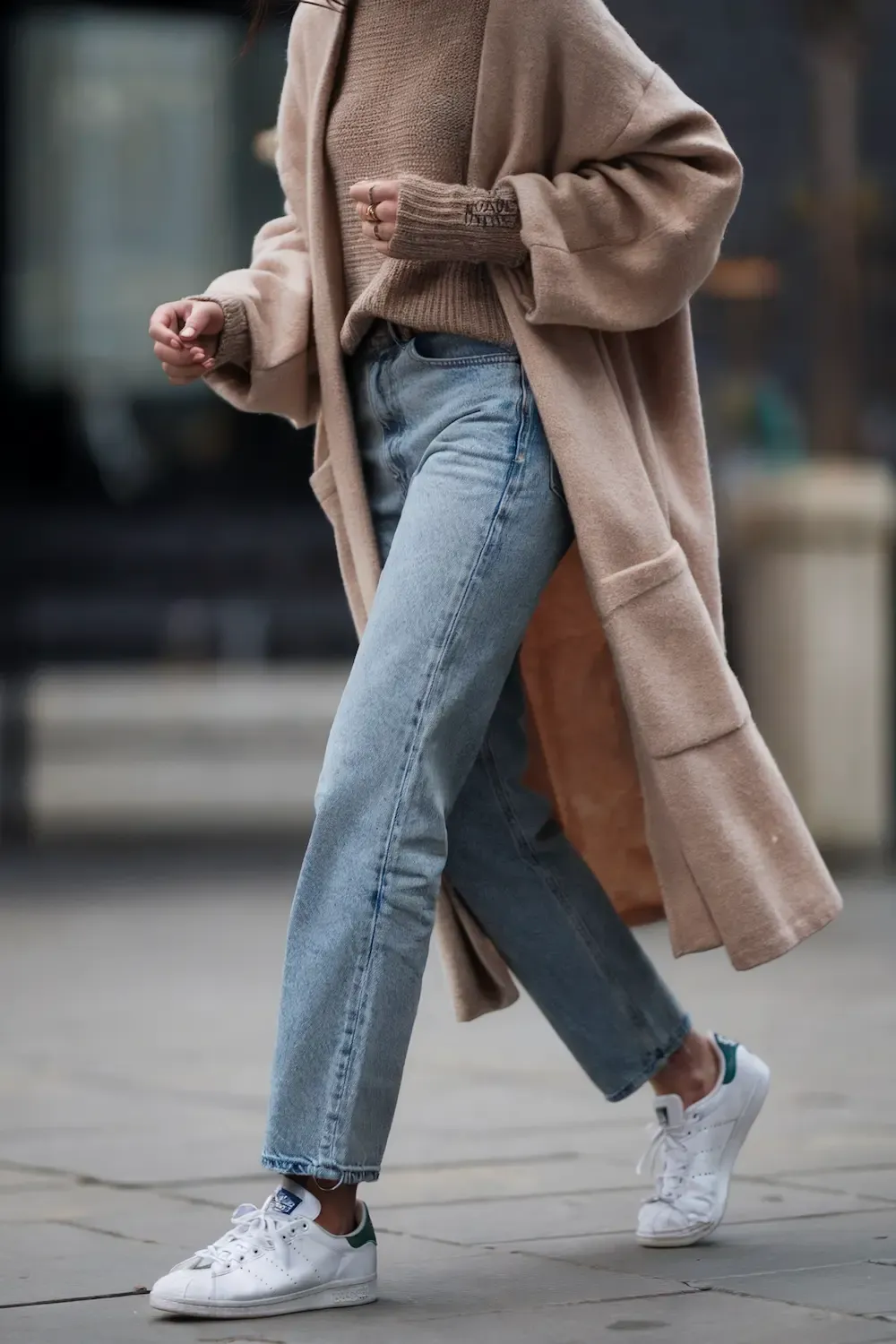 Woman in beige oversized coat, camel sweater, light wash jeans, and white Stan Smith sneakers walking on urban street in fall/winter style