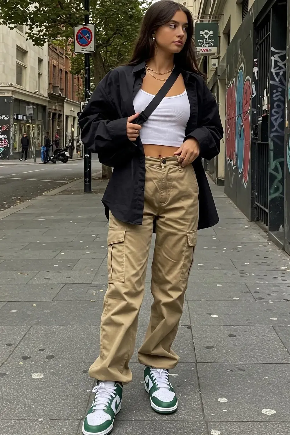 Woman in khaki cargo pants and white/green Nike Dunk Lows, with a white tank and open black shirt, showcasing utilitarian chic.