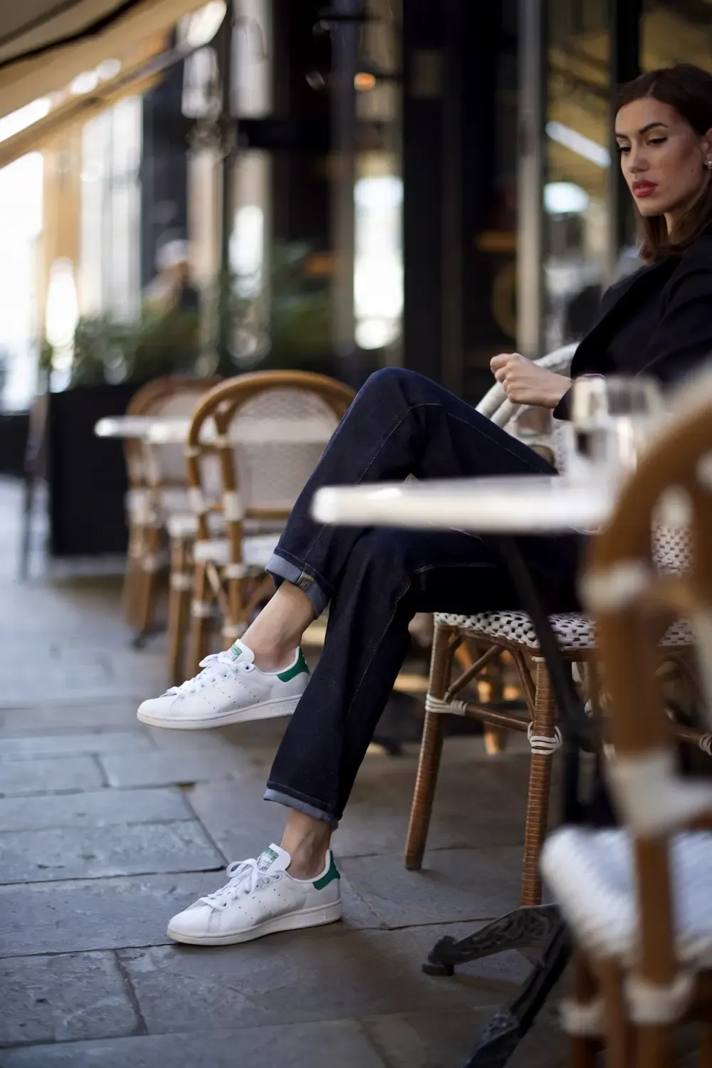 Woman seated at outdoor café wearing dark wash jeans, white Stan Smith sneakers, and dark blazer for a casual evening look