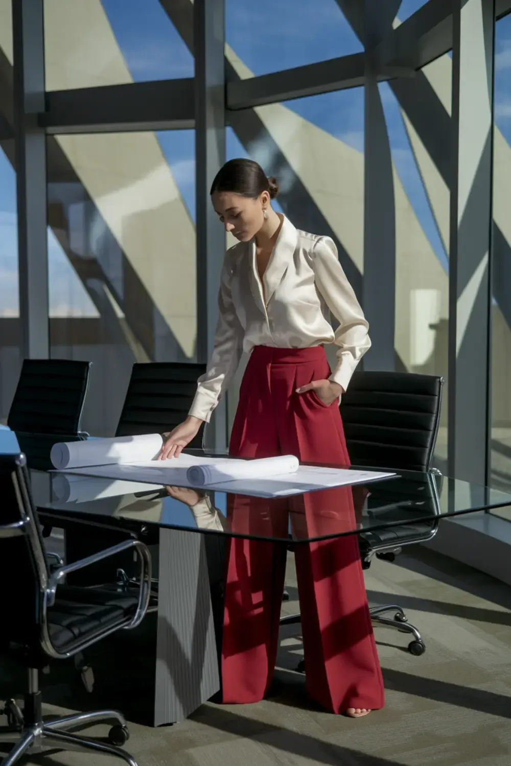 Woman in crisp white blouse and bold red wide-leg trousers reviewing blueprints in modern conference room, paired with neutral New Balance sneakers for comfortable authority.