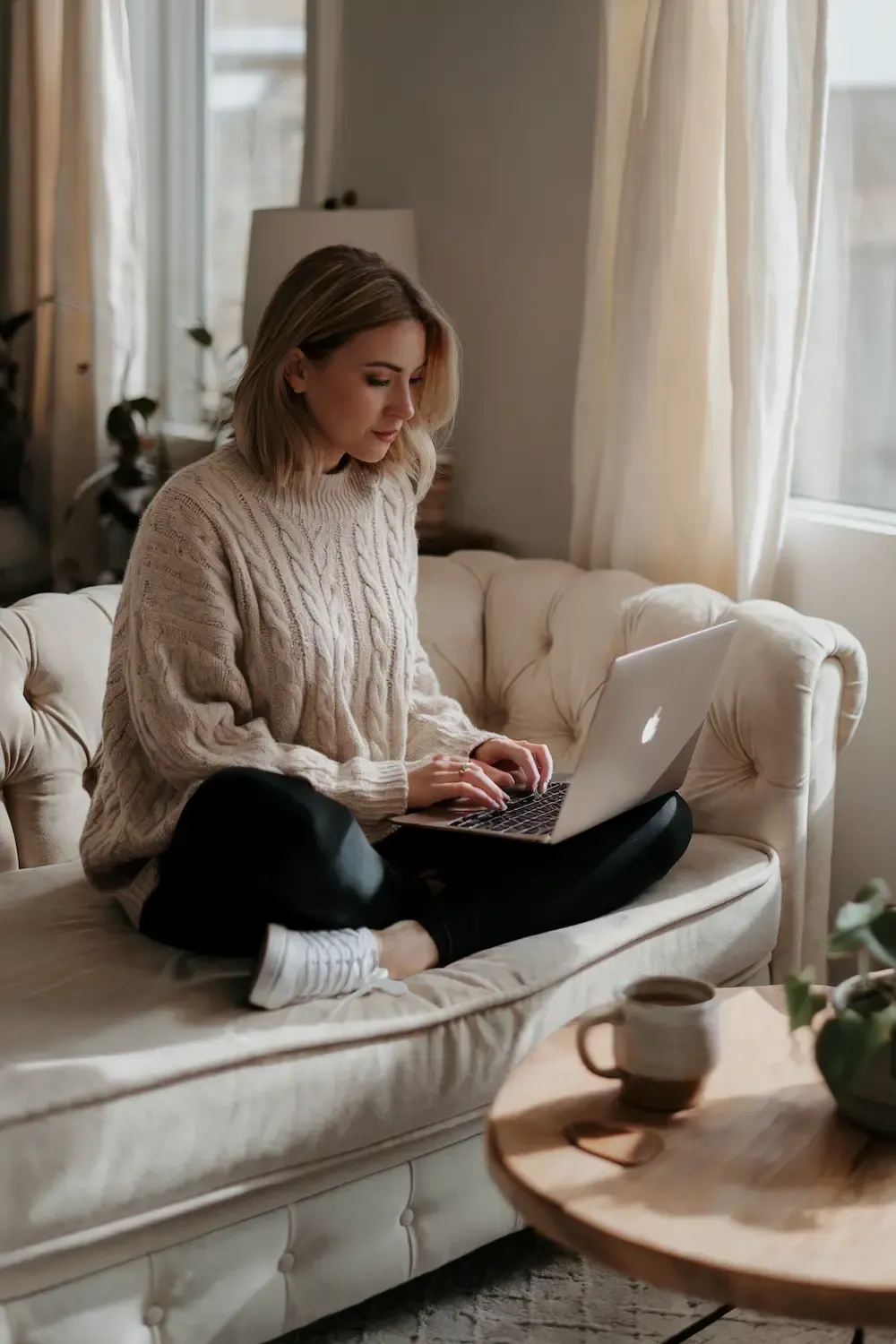 Woman in cream cable-knit sweater and black leggings working on laptop with white sneakers in bright living room - elegant yet comfortable work-from-home outfit for focused productivity