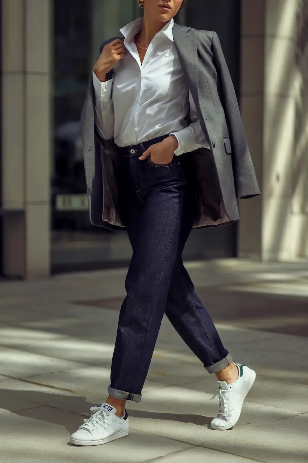Business casual outfit featuring a woman wearing dark raw denim jeans, white button-down shirt, grey blazer, and Stan Smith sneakers