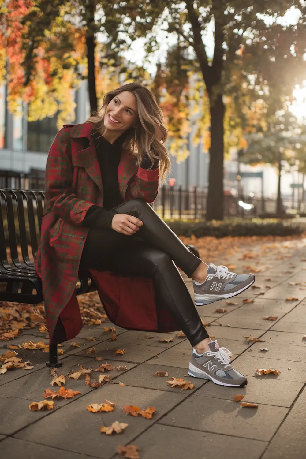 Model wearing red and green plaid midi trench coat with black sweater, leather leggings and gray New Balance 574 sneakers in autumn park setting with golden sunlight, demonstrating seasonal holiday-inspired styling.