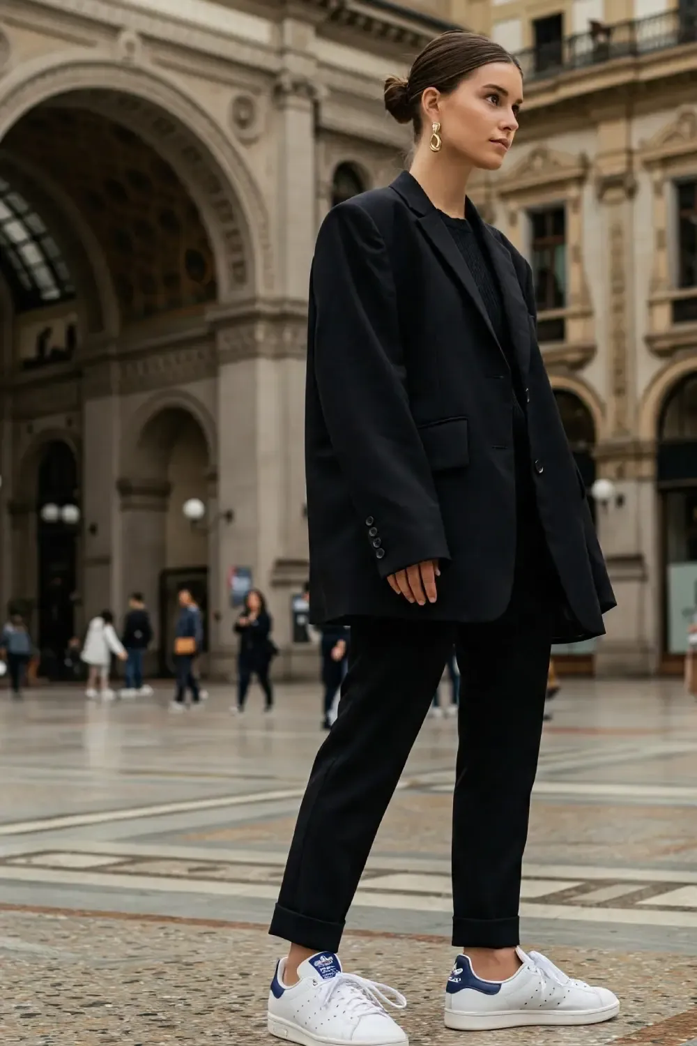 Woman in oversized black blazer and matching trousers outside historic Milan arcade, creating a modern silhouette with sleek white sneakers.