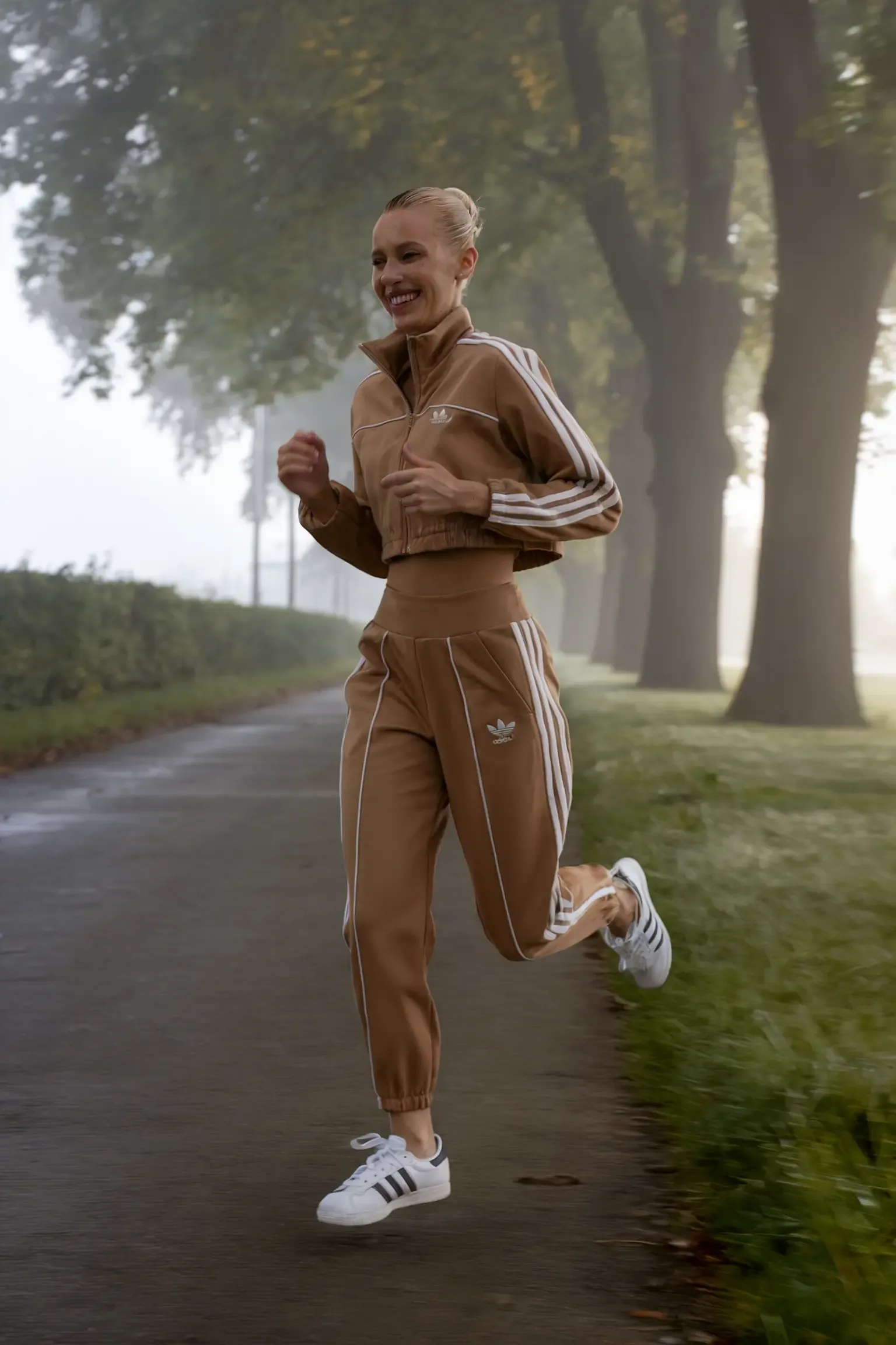 Athletic morning outfit with coordinated tan Adidas tracksuit and white Superstar sneakers, person jogging on misty tree-lined path during sunrise