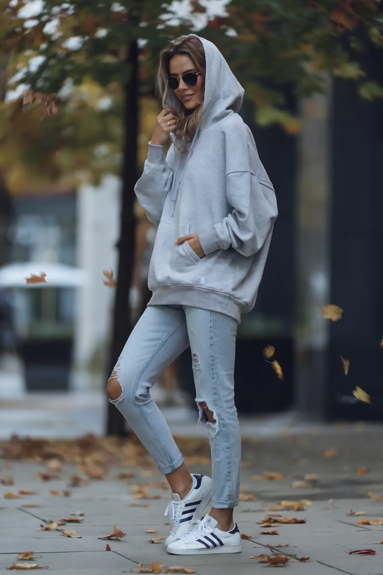 Person in oversized grey hoodie and distressed light wash jeans walking through fall foliage, paired with classic white Adidas sneakers, round sunglasses completing the casual autumn look