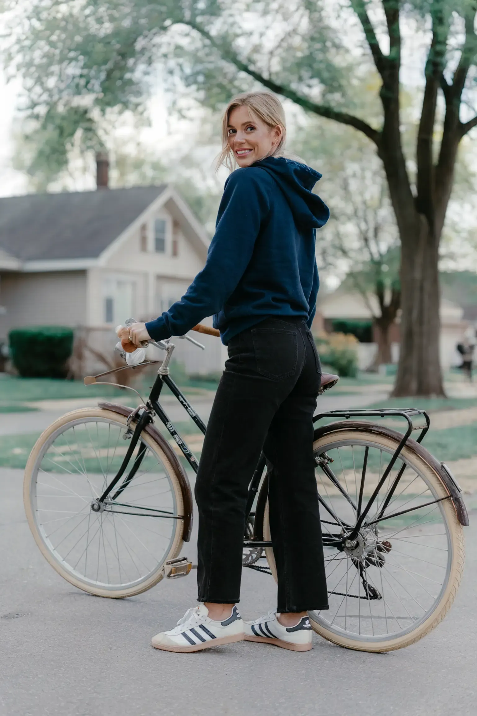 Person with vintage bicycle wearing navy blue hoodie and black straight-leg jeans, paired with white Adidas sneakers, set against suburban residential backdrop