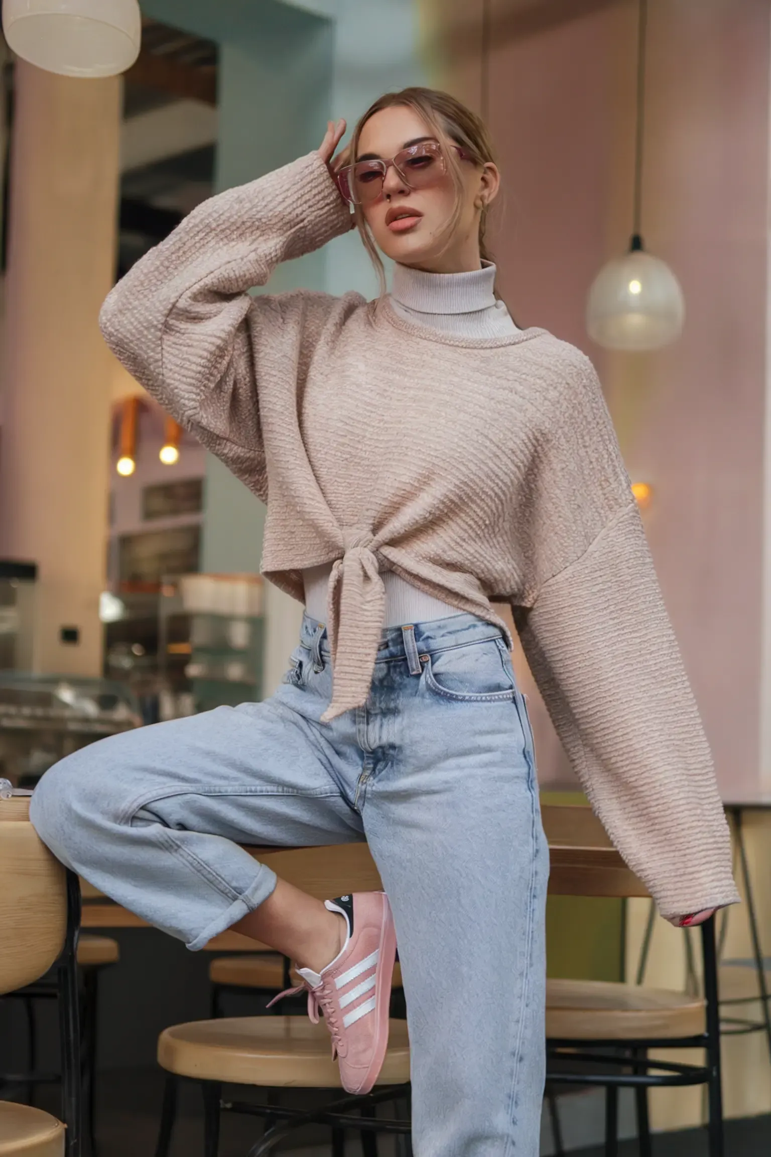 Modern casual look with beige tied sweater over white turtleneck, paired with light wash mom jeans and pink sneakers, photographed in contemporary cafe setting