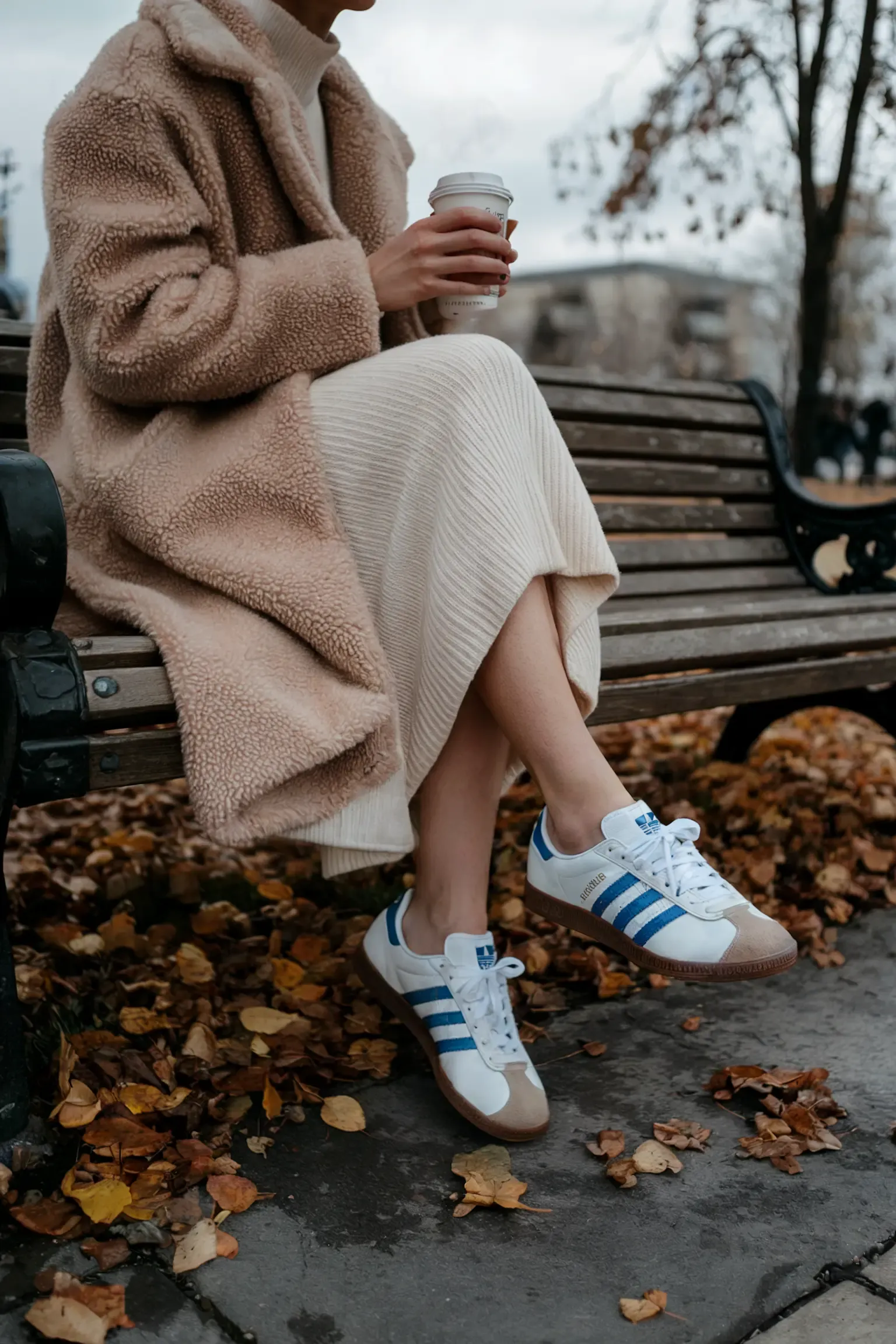 Cozy autumn outfit featuring beige teddy coat, cream ribbed dress and white Adidas Samba sneakers, person sitting on wooden park bench surrounded by fallen leaves holding coffee cup