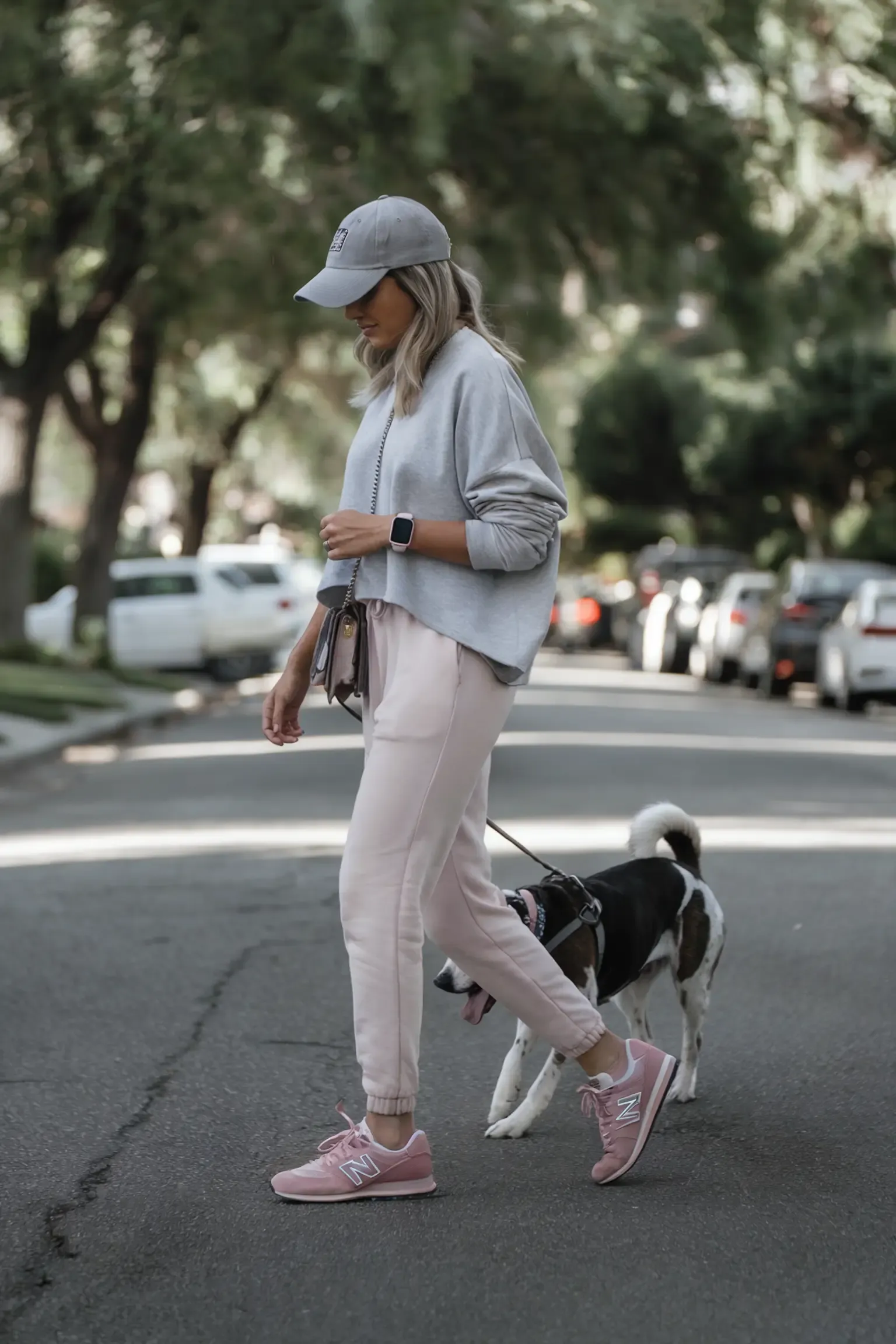 Woman walking dog in light pink joggers and matching New Balance sneakers, paired with oversized grey sweater and baseball cap, showing relaxed weekend athleisure style