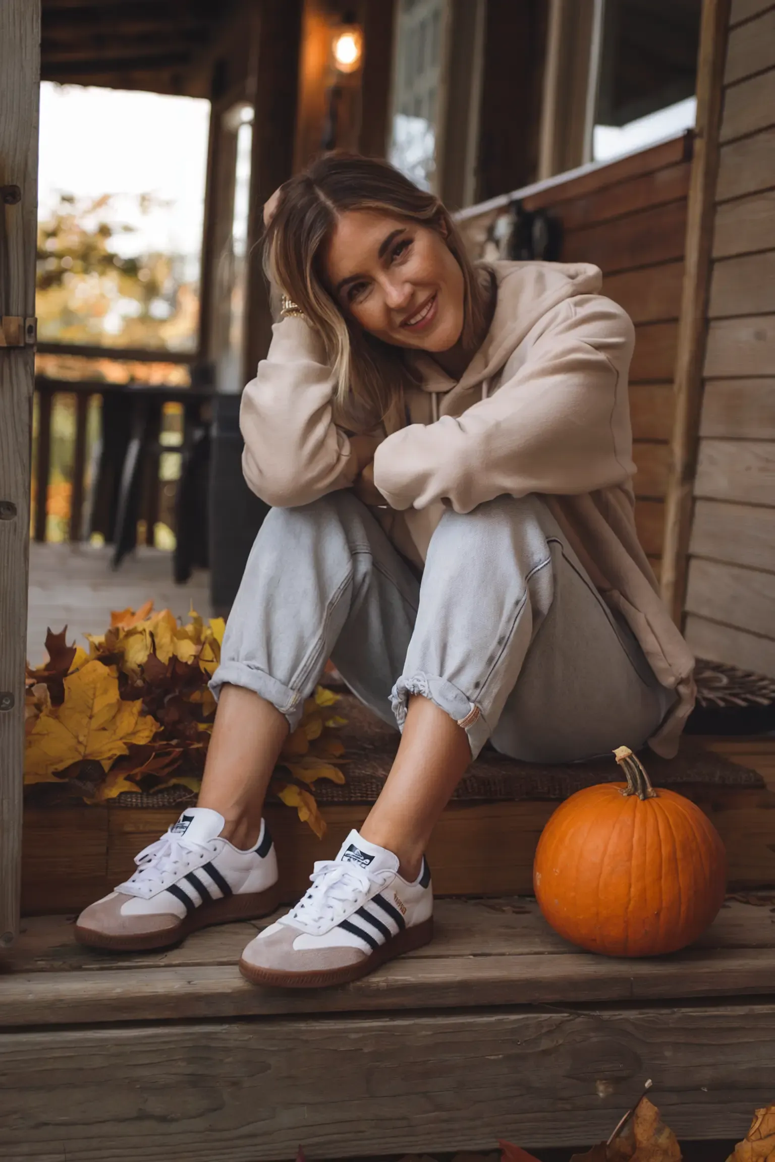Person sitting on wooden porch surrounded by autumn leaves and pumpkin decoration, wearing beige hoodie and light wash jeans with Adidas Samba sneakers, creating a warm seasonal mood
