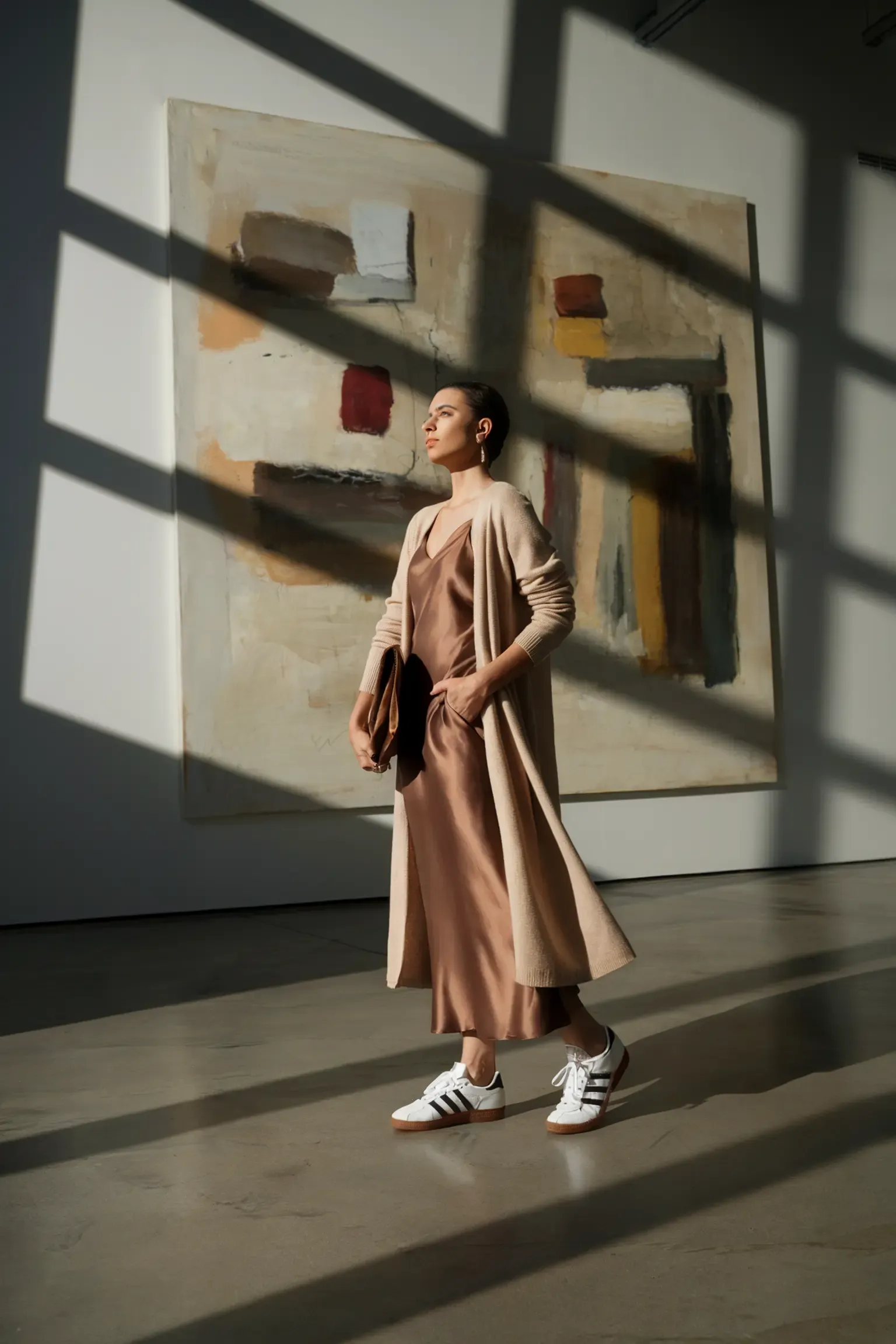 Gallery ready outfit with long beige cardigan, silk slip dress in caramel tone and white Adidas Samba sneakers, person standing against abstract painting with dramatic window shadows