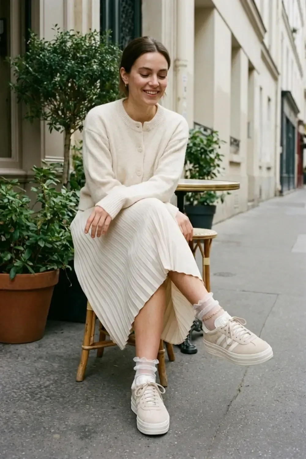  A woman sitting at a Parisian-style café table. She wears a cream pleated midi skirt and a matching cardigan with Adidas Gazelle Bold sneakers