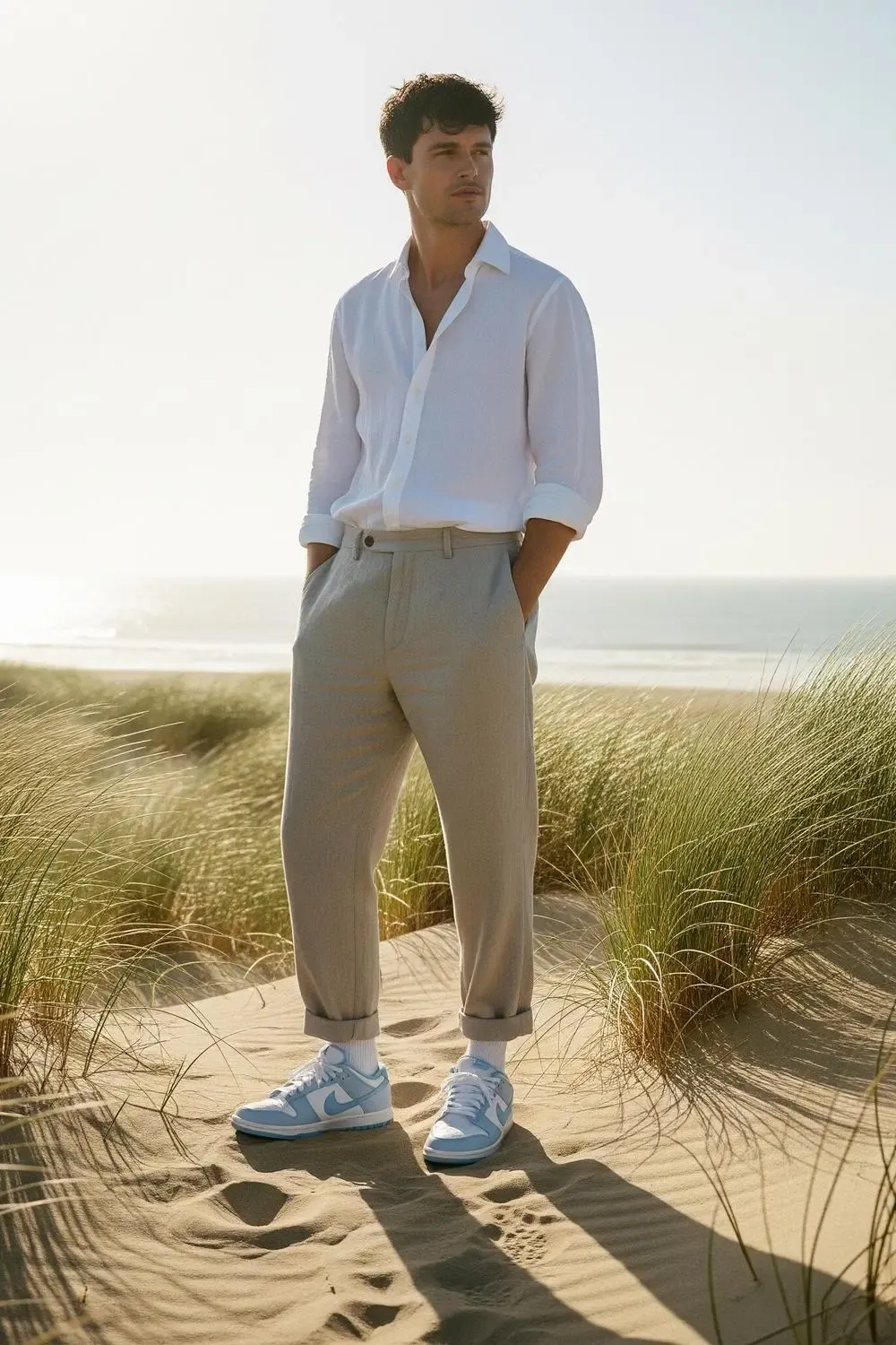 A man casually dressed in beige linen trousers, a white linen shirt partially unbuttoned, and Nike Dunk Low 'UNC' sneakers, standing on a sandy dune with the ocean in the background.