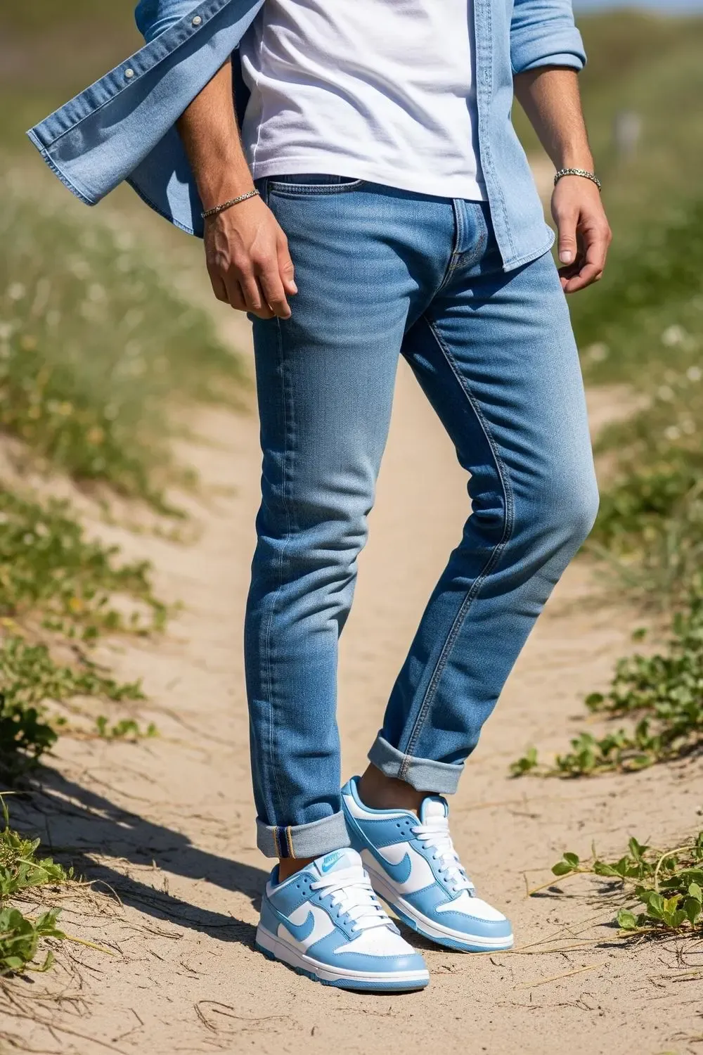 A man embodying a relaxed coastal weekend style, wearing light-wash cuffed jeans, a white t-shirt, an open light blue chambray shirt, and Nike Dunk Low 'UNC' sneakers, seen from mid-thigh up, walking along a sun-dappled beach path.
