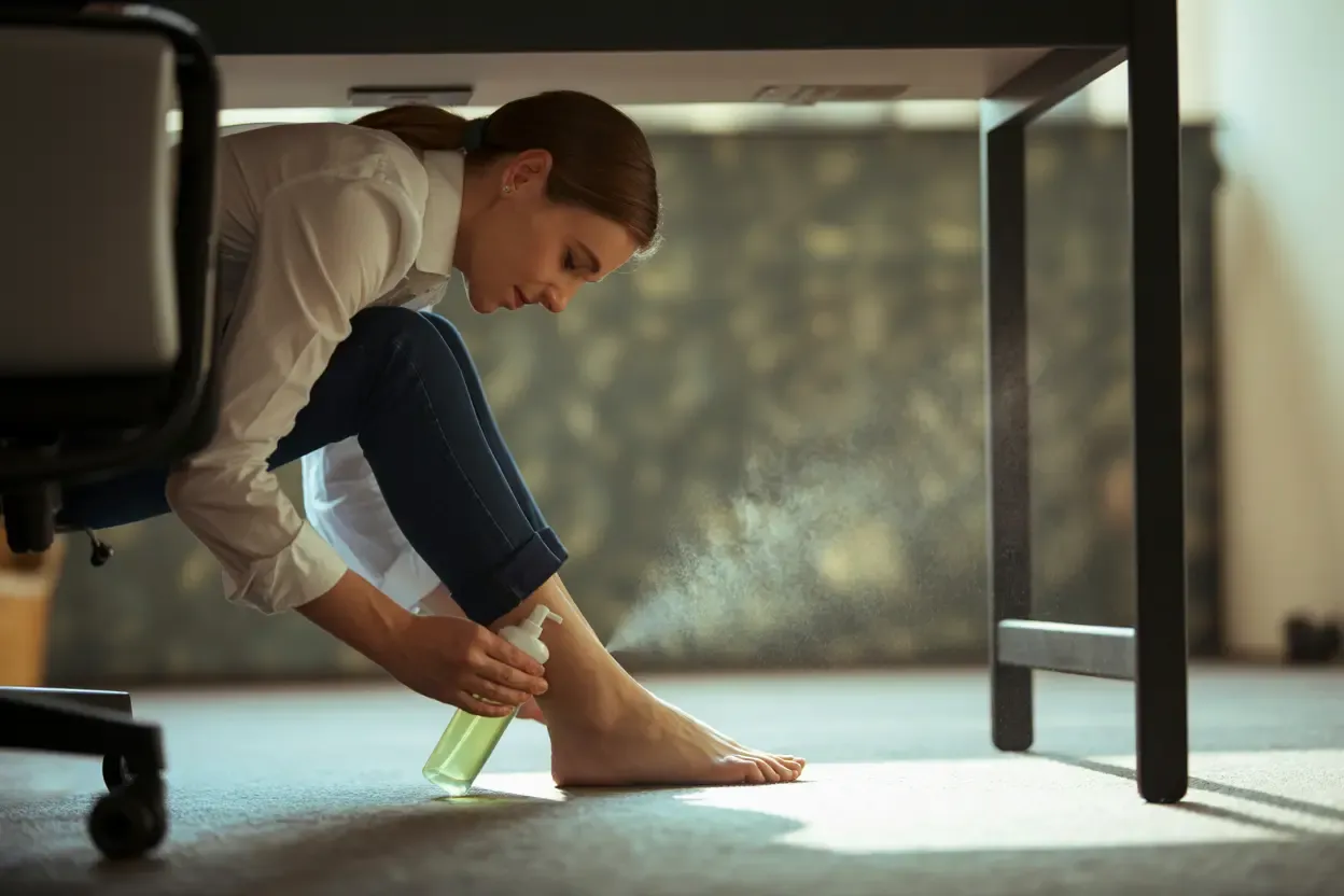Image showing a person discreetly using a foot spray or wipe on their foot, illustrating a mid-day refreshment technique.
