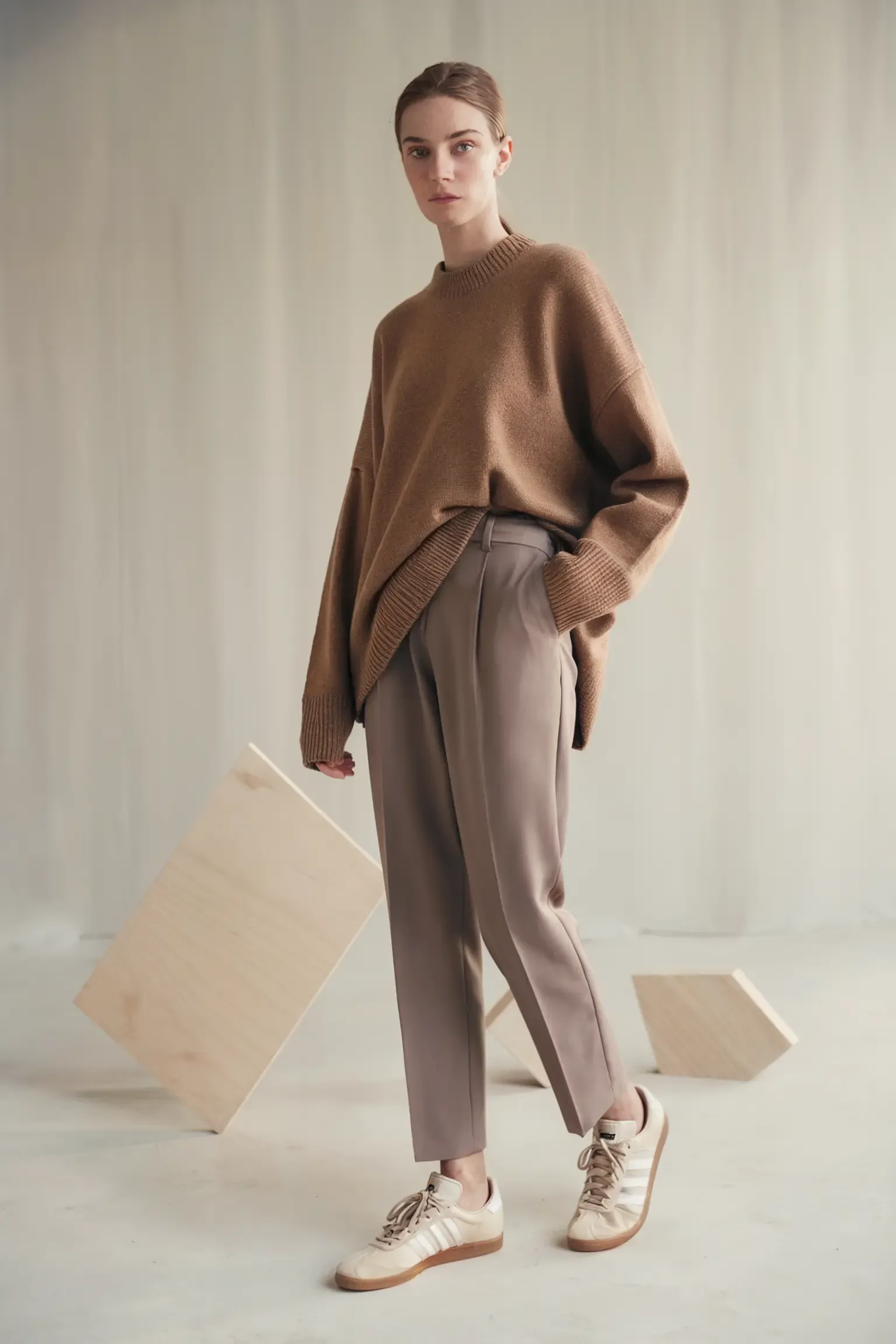 Minimalist outfit featuring an oversized camel sweater paired with tapered beige trousers and cream sneakers, photographed in a studio setting with geometric props