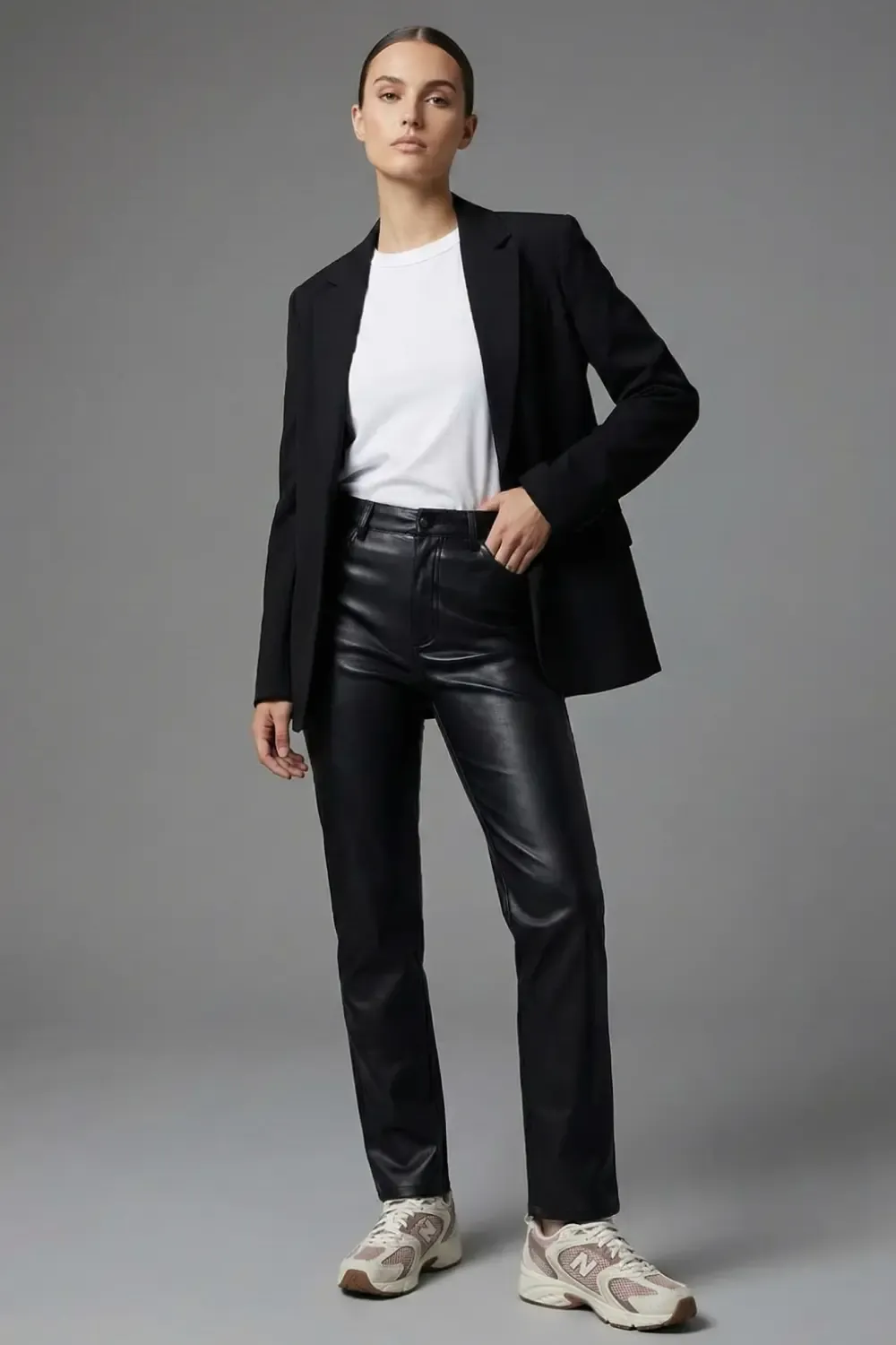 Woman wearing a black blazer, white T-shirt, leather pants, and New Balance 530 sneakers in a studio setting.