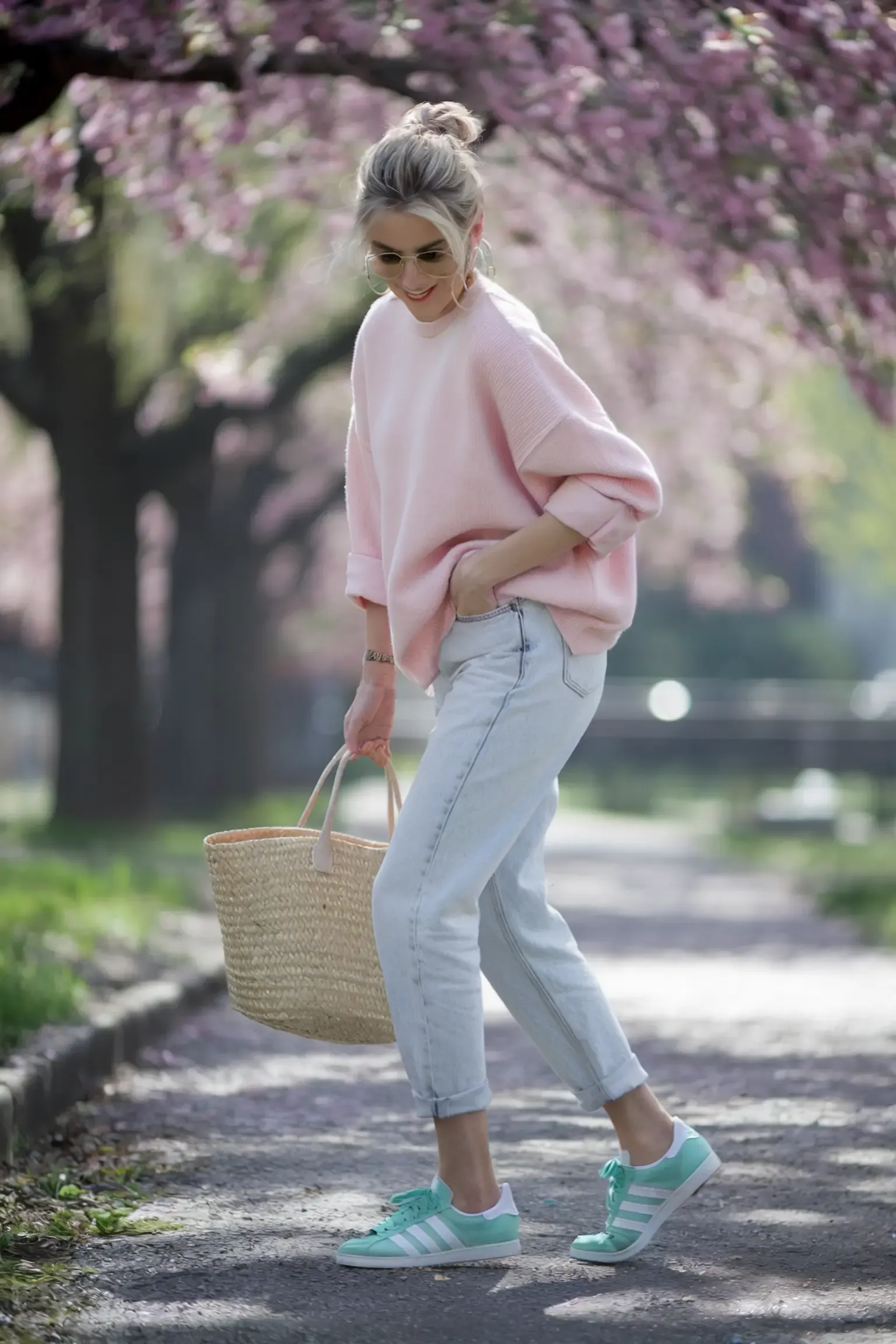 Casual spring outfit with oversized pink sweater and light wash mom jeans, accessorized with straw market bag, photographed under blooming cherry blossoms