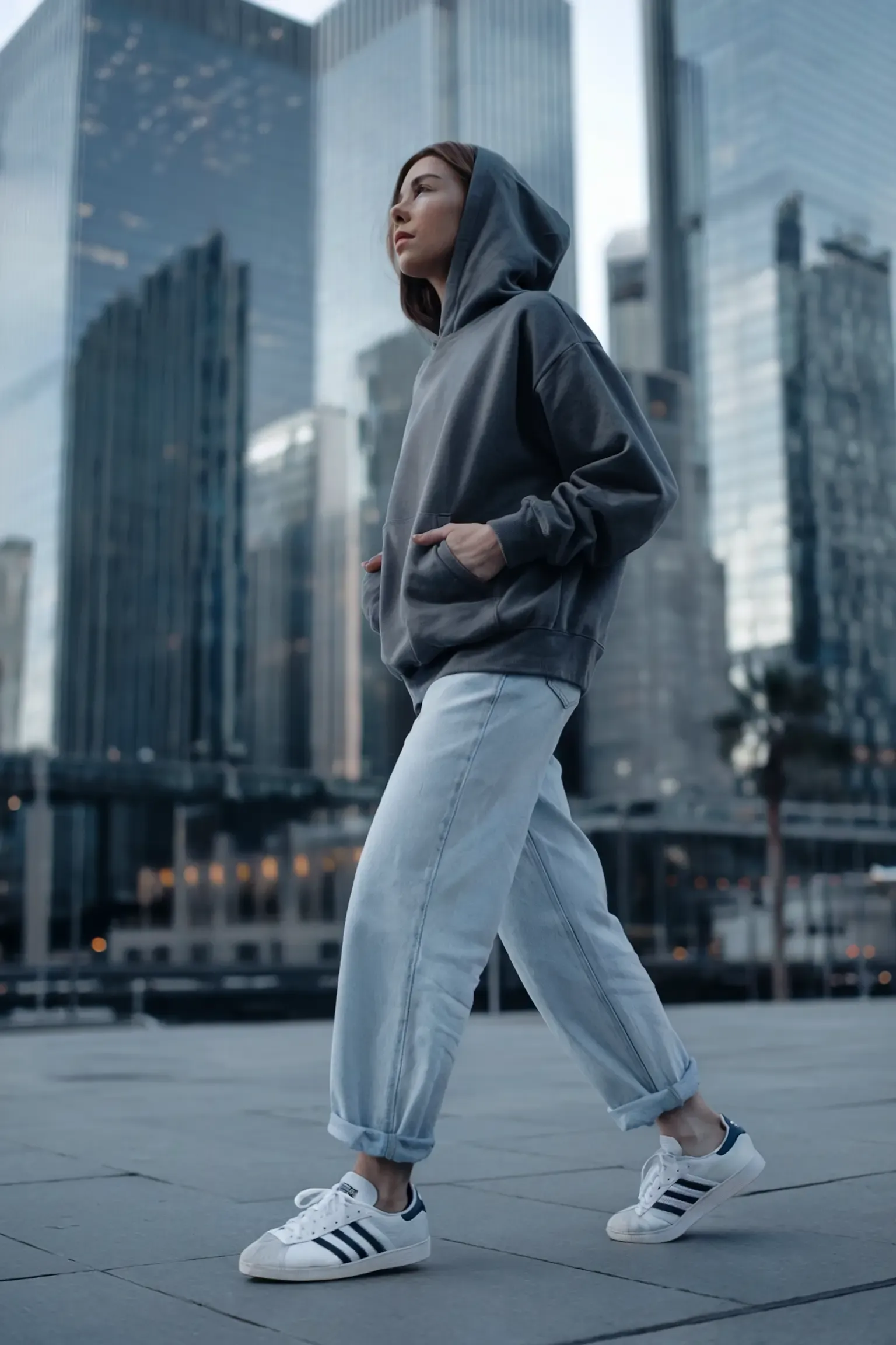 Person wearing charcoal grey hoodie and light wash straight-leg jeans against modern glass skyscrapers at dusk, styled with white Adidas sneakers for a clean urban aesthetic