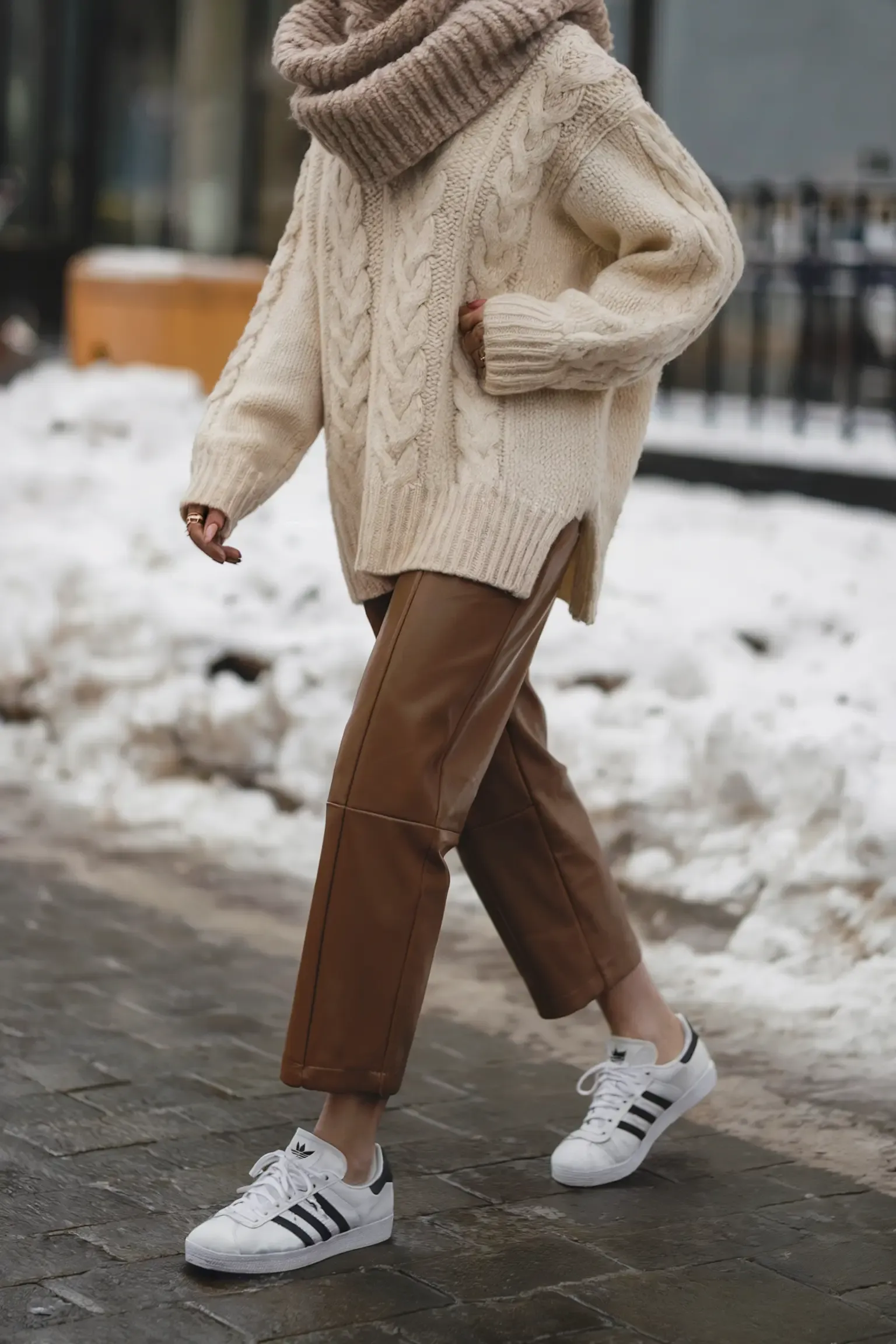 Winter street style featuring cream cable-knit turtleneck sweater with brown leather pants and classic white sneakers, photographed in snowy urban setting
