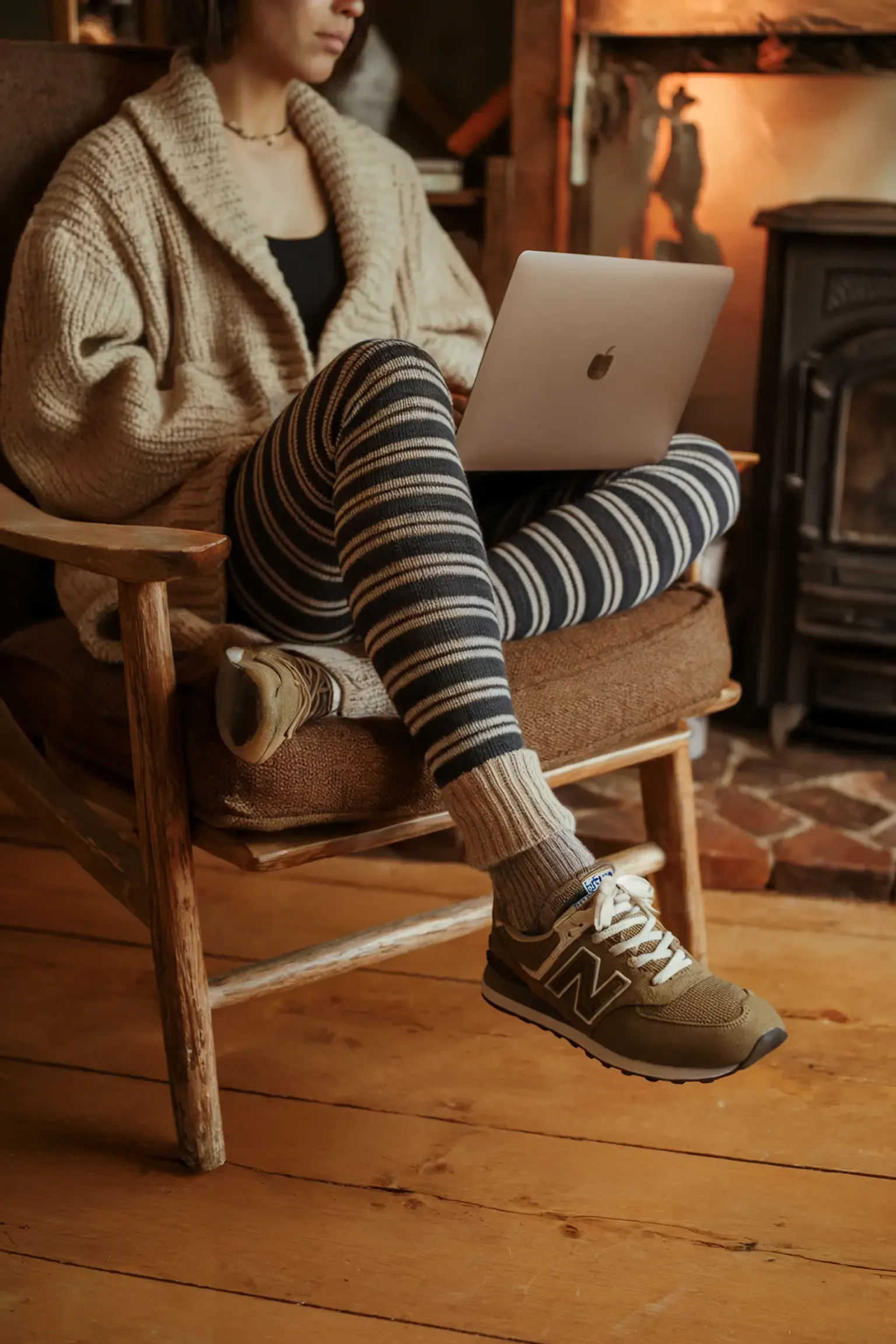 Woman working on laptop in rustic setting wearing chunky beige cardigan and striped leggings, paired with tan New Balance sneakers, highlighting comfortable work-from-home winter style
